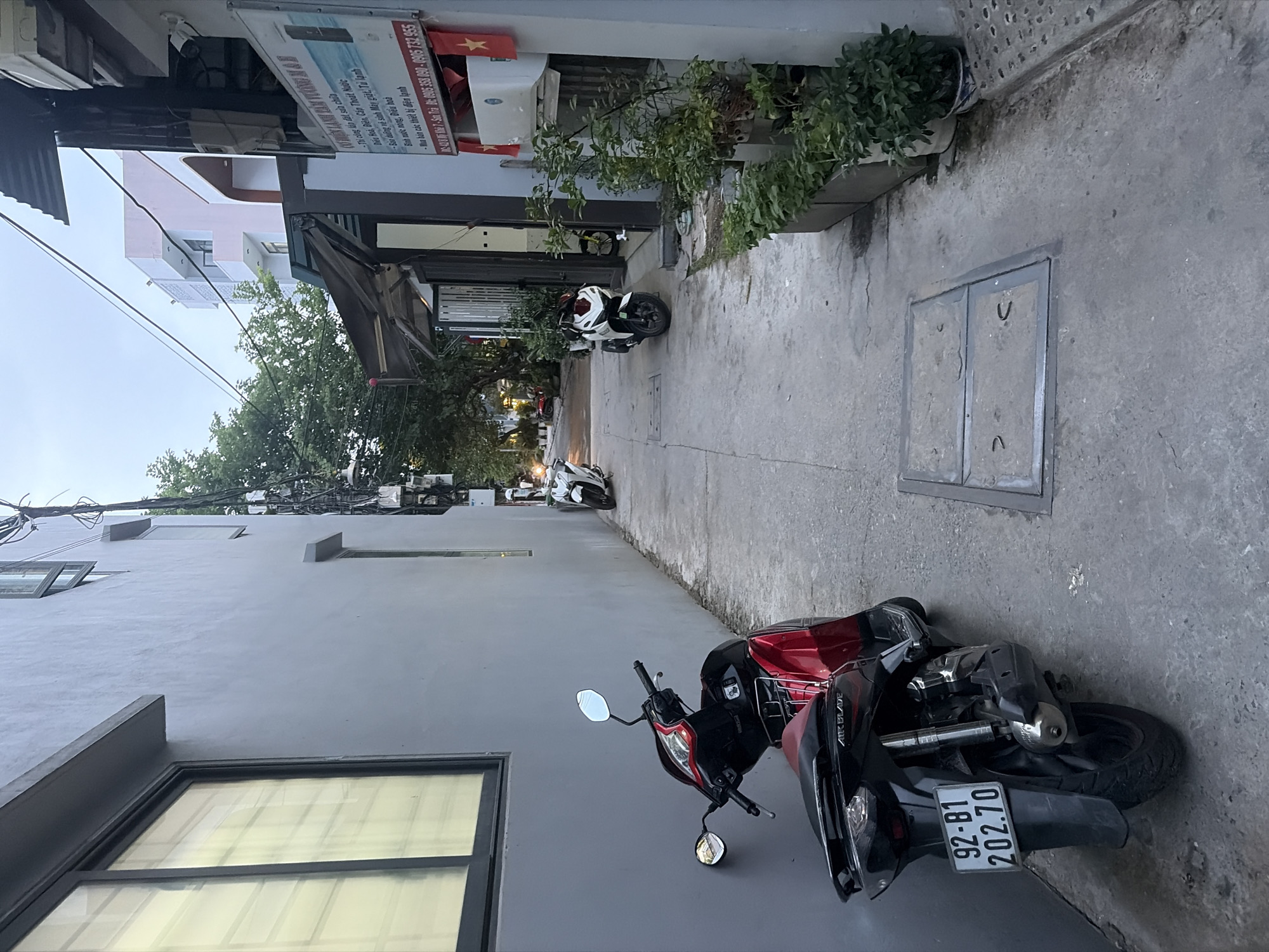 The image depicts a narrow alleyway in the city of Đà Nẵng, Vietnam. On the left, a light gray building with a large, dark-framed window dominates the foreground. To the right, a red and black scooter is parked. Further down the alley, two white scooters are visible. The alleyway is paved with concrete, and there are potted plants and a drain cover on the right side. A sign with Vietnamese text is affixed to the wall of a building on the right. Overhead, power lines are strung across the sky. The lighting suggests it is either early morning or late afternoon.