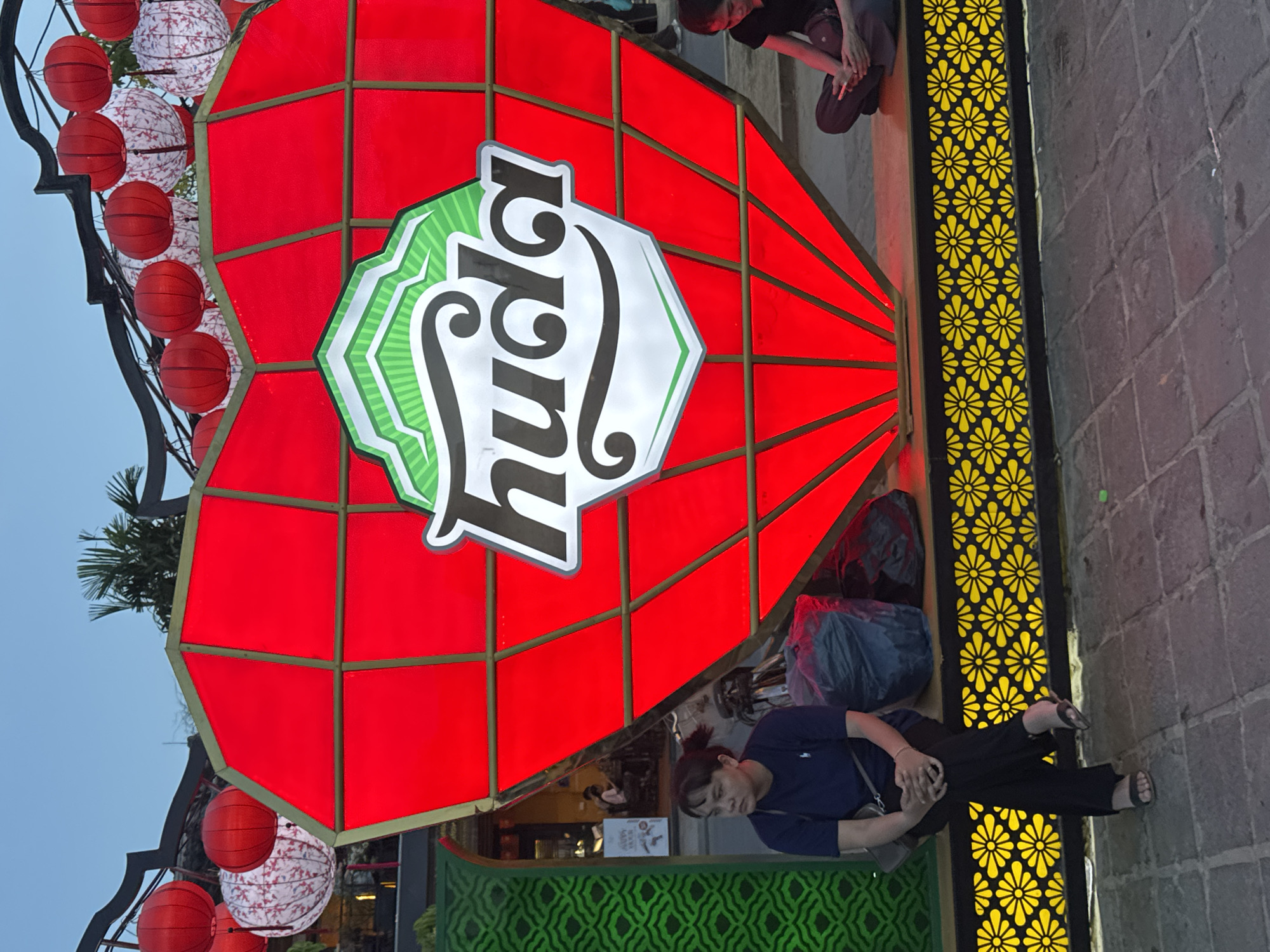 A daytime, ground-level shot, taken in Hội An Ward, Vietnam, presents a busy street scene. Dominating the image is a large, red, heart-shaped advertisement sign for "huda" beer. The sign is adorned with traditional red lanterns on its left side and sits above a building with green patterned decor.
In the foreground, a woman dressed in a blue shirt and black pants stands with her arms crossed. Nearby, black and yellow floral patterns decorate the surrounding structures.
The background features a mix of traditional and modern architecture, capturing the dynamic atmosphere of Hội An. The sky is partly visible, offering a glimpse of a clear day.