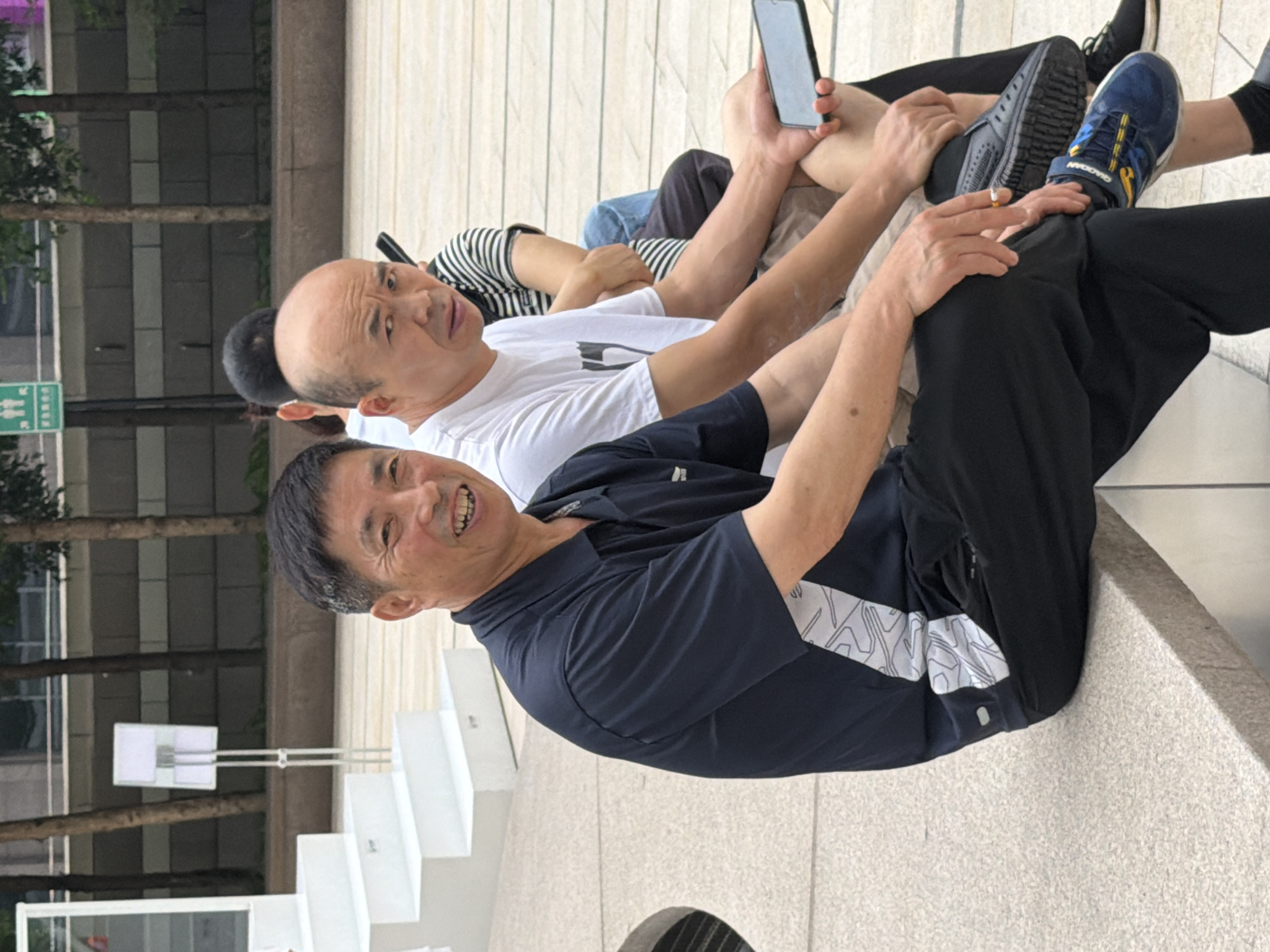 In Yuzhong District, China, three men are seated on a textured, light-colored outdoor bench. The man on the left, facing the camera, has short dark hair and is wearing a dark blue polo shirt with a white geometric pattern on the sleeve and black pants. He is smiling. To his right, a bald man is looking to his left with a neutral expression and is wearing a white t-shirt. Behind him, a man with dark hair is visible, wearing a striped t-shirt and dark pants. The man on the left is holding a lit cigarette in his right hand. The bald man on the right is holding a dark-colored smartphone in his right hand. The background includes trees, a building with windows, and a white tiered structure. The ground is paved with light-colored tiles.