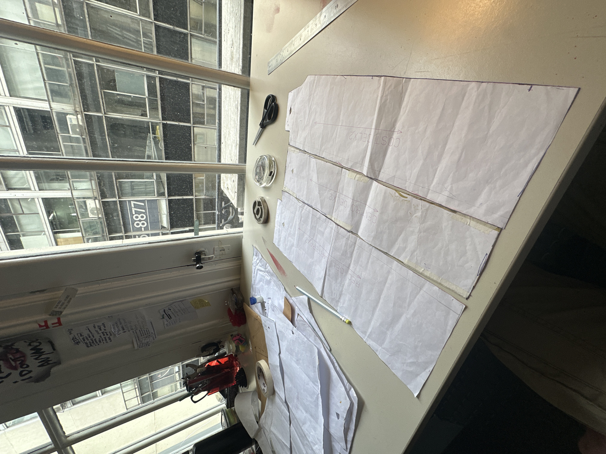 The image depicts a cluttered workspace, likely indoors, with a focus on sewing-related objects. A window shows a view of a building across the street, with the number "-8877" partially visible on the building facade. Sewing patterns are laid out on a table, along with scissors, bobbins, a ruler, and other sewing tools. Other papers and adhesives are visible near the window. The scene suggests a sewing project in progress. No people are directly visible in the image. It is not possible to identify the location within Buenos Aires, Argentina solely based on the given information.