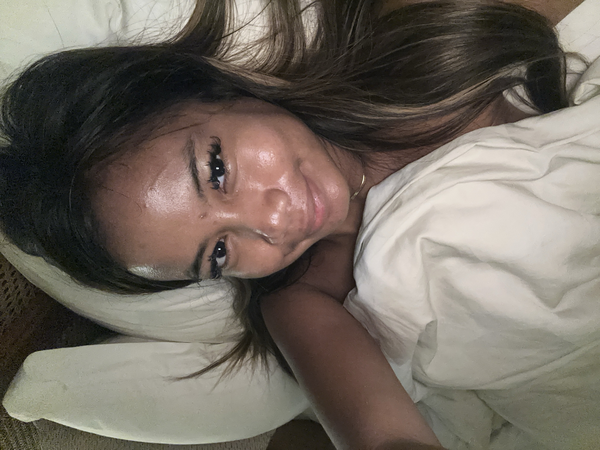The image is a close-up selfie of a woman lying in bed. She is mostly covered by a white sheet or blanket, with only her face and one arm visible. Her hair is dark brown with lighter highlights and is partially spread out on the pillow. She has dark eyes and appears to be wearing false eyelashes. A thin gold necklace is visible around her neck. The background suggests a bedroom setting with a pillow and bedding. There is no discernible information to confirm it is located in Ubud District, Indonesia.