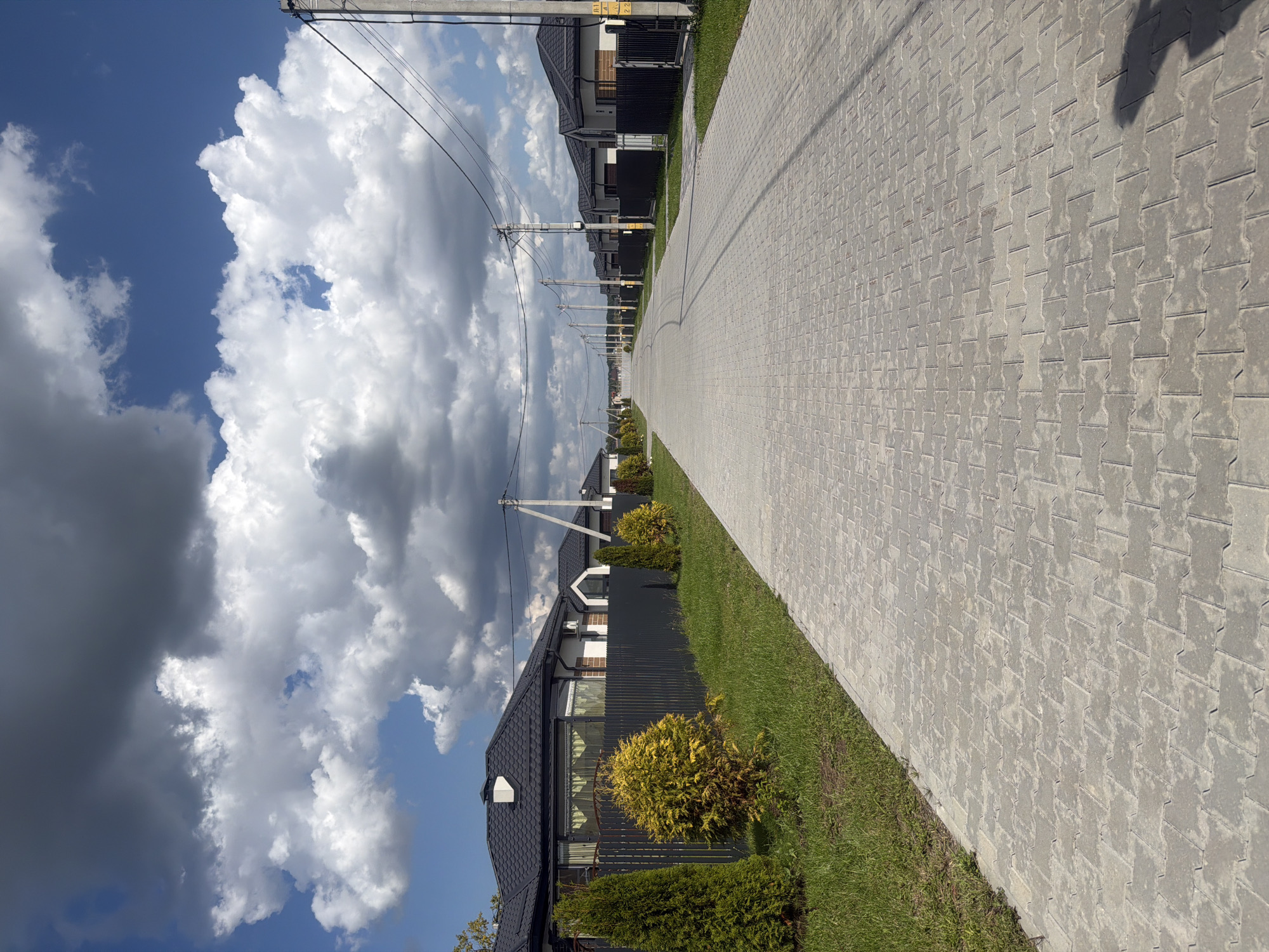 The image depicts a street in the city of Golubevo, Russia. On the left side of the street, there are several houses with dark roofs and black fences. Some houses have manicured lawns with evergreen trees and shrubs. The street itself is paved with interlocking concrete pavers, and it extends into the distance. On the right side of the street, there are more houses with similar architectural features, and telephone poles with power lines are visible along the street. The sky is partly cloudy with dramatic cumulus clouds and patches of blue sky. The overall scene suggests a suburban residential area with modern construction.