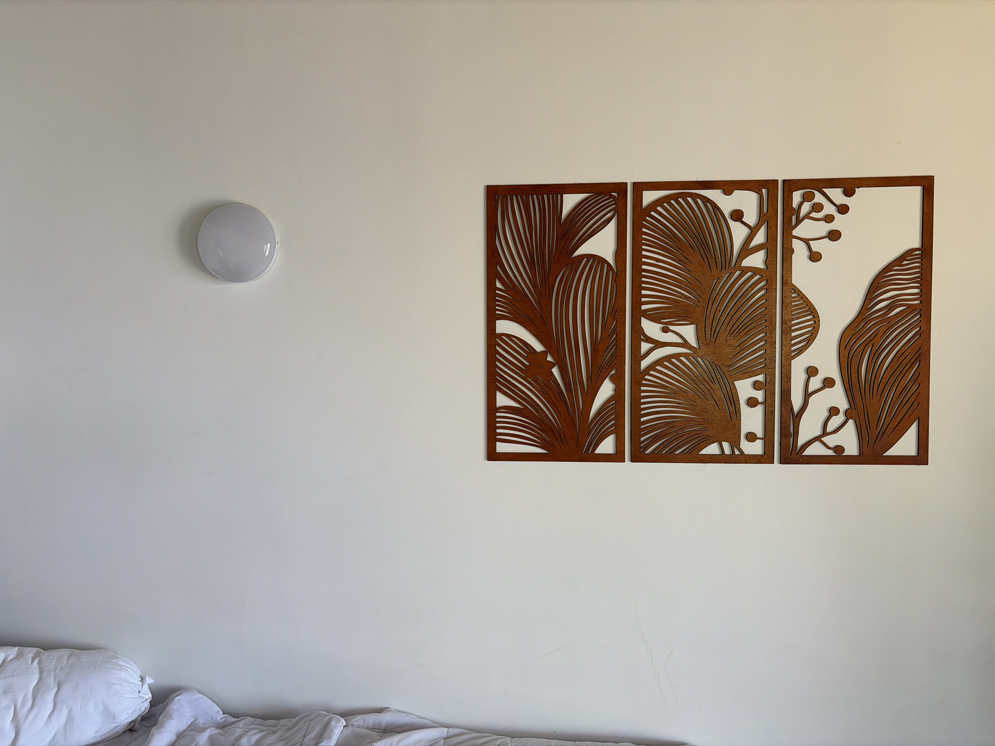 In Abang, Indonesia, a plain white wall serves as the backdrop for a contemporary bedroom. To the left, a circular, white ceiling light fixture is mounted. To the right, a triptych of decorative wall art, featuring intricate brown leaf and branch patterns within square frames, is displayed. In the foreground, the edge of a bed with white linens and a large pillow is visible, suggesting a domestic setting. The lighting in the image is soft, possibly from natural sunlight, casting gentle shadows.