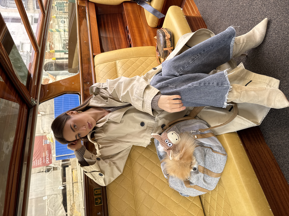 A female individual is seated on a yellow quilted leather bench inside a polished wooden water taxi in Venice, Italy. She has long dark hair, is wearing a light beige trench coat over a grey ribbed top, blue raw-hem jeans, and light beige pointed-toe ankle boots. Gold hoop earrings are visible. Her right hand is resting on her head, and her left hand is on her lap. She looks directly at the camera with a neutral expression. To her left, a grey duffle bag with brown leather accents contains a small, fluffy light brown dog, with a brown knitted animal charm attached to the bag. To her right, on the same bench, rests a small brown leather-trimmed trunk-style bag. Through the boat's windows, blurred Venetian architecture is visible, along with some construction scaffolding or blue tarpaulins in the background, indicative of an urban canal environment.