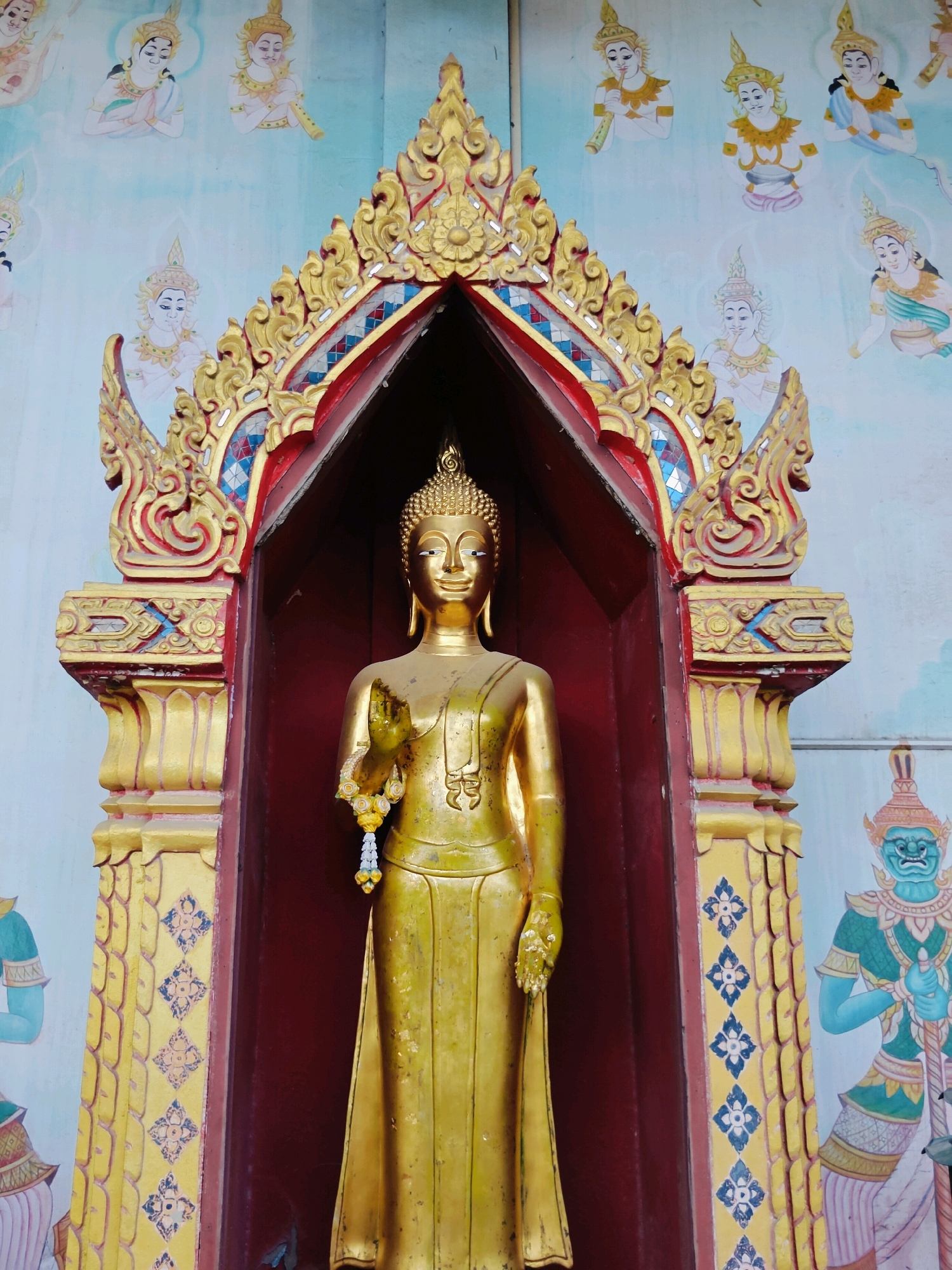 A full-shot, eye-level, front view photograph captures a golden Buddha statue positioned centrally within a shrine. The statue is adorned with a garland of white and yellow flowers draped over its right shoulder and hand. The shrine features ornate golden architectural elements, including a tiered, decorative archway above the Buddha, with red and blue accents. The background wall is adorned with a mural depicting figures in traditional attire and a blue-skinned guardian. The image is taken in Phra Nakhon Si Ayutthaya City Municipality, Thailand.