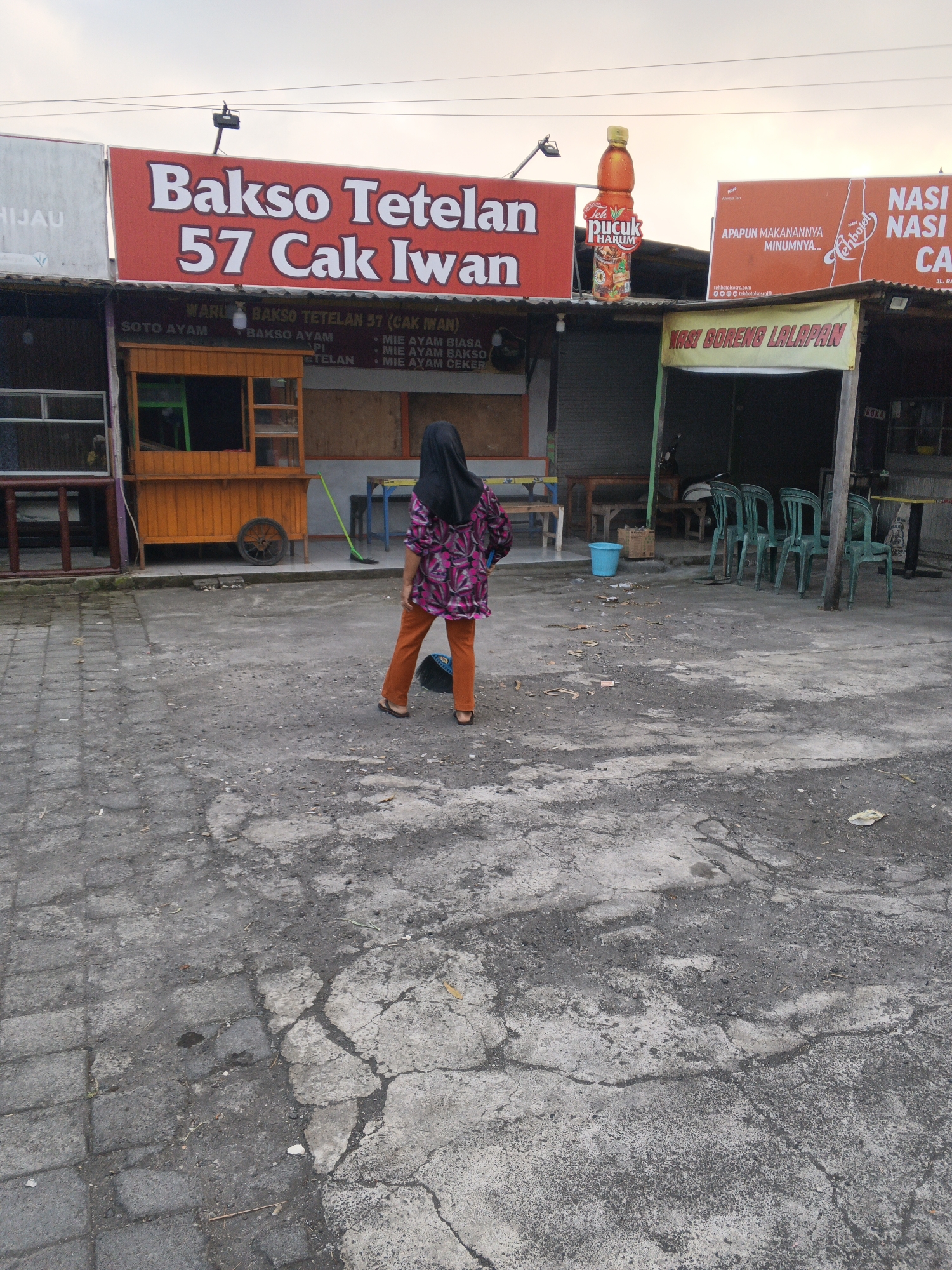 A full shot shows a Muslim woman wearing an orange headscarf and a pink and purple patterned shirt walking away from the camera. She is wearing orange pants and flip-flops. She is standing in front of a food stall called "Bakso Tetelan 57 Cak Iwan" in Sukawati, Indonesia. In the background, there are green plastic chairs and tables outside the stall. The ground is made of cracked concrete and cobblestones. The sky is overcast.