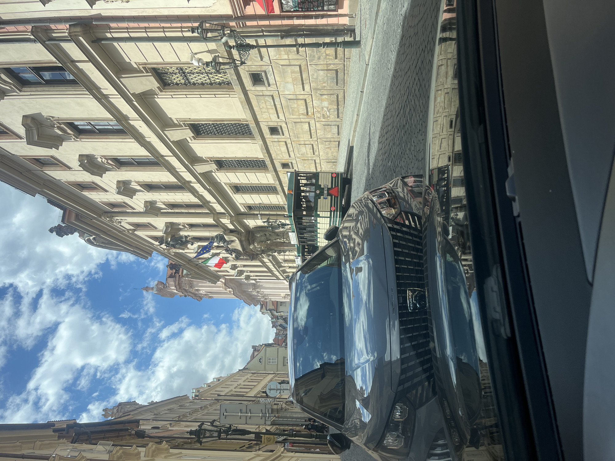 The image depicts a street scene in Prague, Czechia, likely in the Old Town or Lesser Town due to the cobblestone street and baroque architecture. In the foreground, the hood and windshield of a dark grey Peugeot 2008 are visible, reflecting the surrounding buildings and sky. Ahead, a green and cream-colored tourist tram travels down the street, carrying passengers. The tram is positioned on the right side of the road. On the left and right sides of the street, ornate buildings with classical architectural elements line the narrow lane. The sky is partly cloudy, with patches of blue sky visible between white clouds. The overall atmosphere suggests a sunny day in a historic European city.