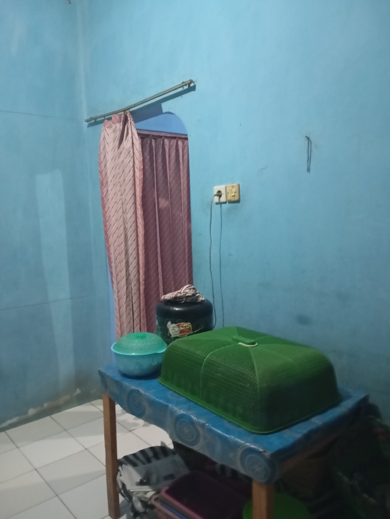 The image depicts an indoor scene, likely a room in a residence in Kalitinggar, Indonesia. The walls are painted a light blue color. In the background, a pink curtain is visible, partially obscuring a doorway or window. A metal rod runs horizontally across the top portion of the wall near the curtain, likely serving as a curtain rod.
In the foreground, a table with a blue patterned cloth covers its surface. On the table, there are several objects. A teal-colored bowl with a lid is placed on the left side of the table. To its right, a large black container with a patterned cloth on top is situated. In front of this container is a large, green, dome-shaped food cover. Below the table, various items are visible, including what appears to be bags or storage containers with striped patterns. A wooden leg of the table is also visible.
On the blue wall, towards the right side of the image, an electrical outlet with two sockets is mounted. A black cord extends downwards from the outlet. There is also a vertical element embedded in the wall above and to the right of the outlet. The floor is tiled with white square tiles. The overall impression is that of a functional, albeit simply furnished, living space.