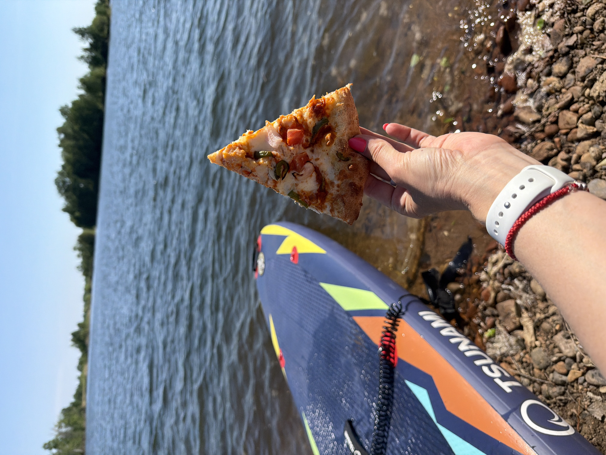 The photo, taken near Razdolnoye, Russia, shows a person holding a slice of pizza in their right hand, with a stand-up paddleboard (SUP) nearby. The pizza has a visible crust and toppings including what appears to be cheese, tomatoes, peppers, and some kind of meat. The hand holding the pizza has manicured nails with pink polish. The person is wearing a white smartwatch and a red bracelet. The SUP is blue with colorful geometric designs and the word "TSUNAMI" printed on it. In the background, there's a body of water, likely a lake or a wide river, with trees visible on the opposite shore. The shoreline where the photo is taken is rocky. The weather appears sunny.