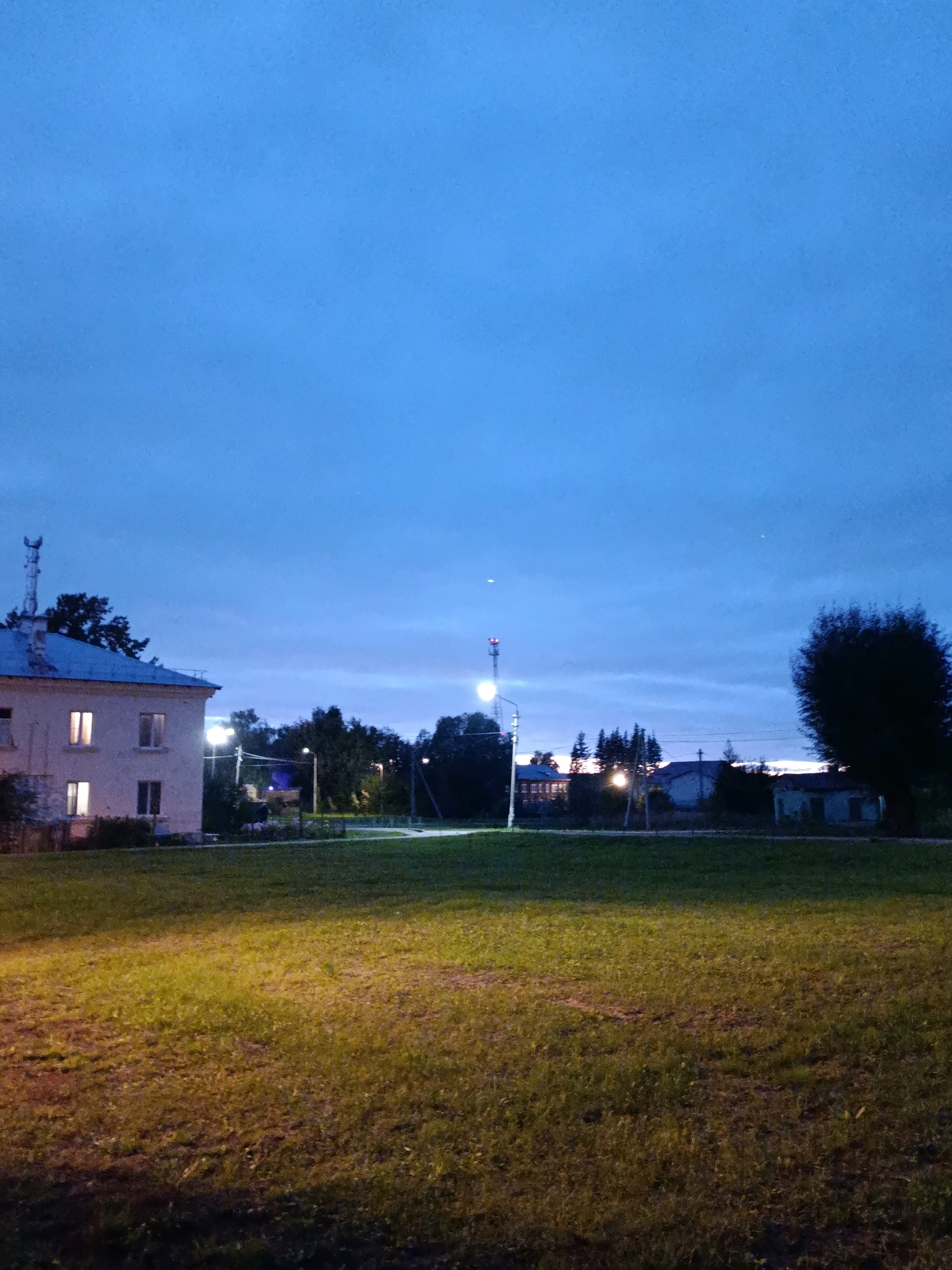 The image depicts a dusky scene in the city of Vereya, Russia. In the foreground, a large expanse of mowed grass is illuminated by artificial light sources. In the middle ground, a light-colored, two-story building with illuminated windows is visible to the left, partially obscured by trees. A streetlamp with multiple lights stands in front of the building. Further into the background, more trees and several other buildings are scattered across the landscape. A tall, slender tower with lights is visible in the distance. The sky is a deep blue, transitioning to lighter shades near the horizon, with a few wispy clouds. The overall impression is of a quiet, semi-urban environment at dusk.