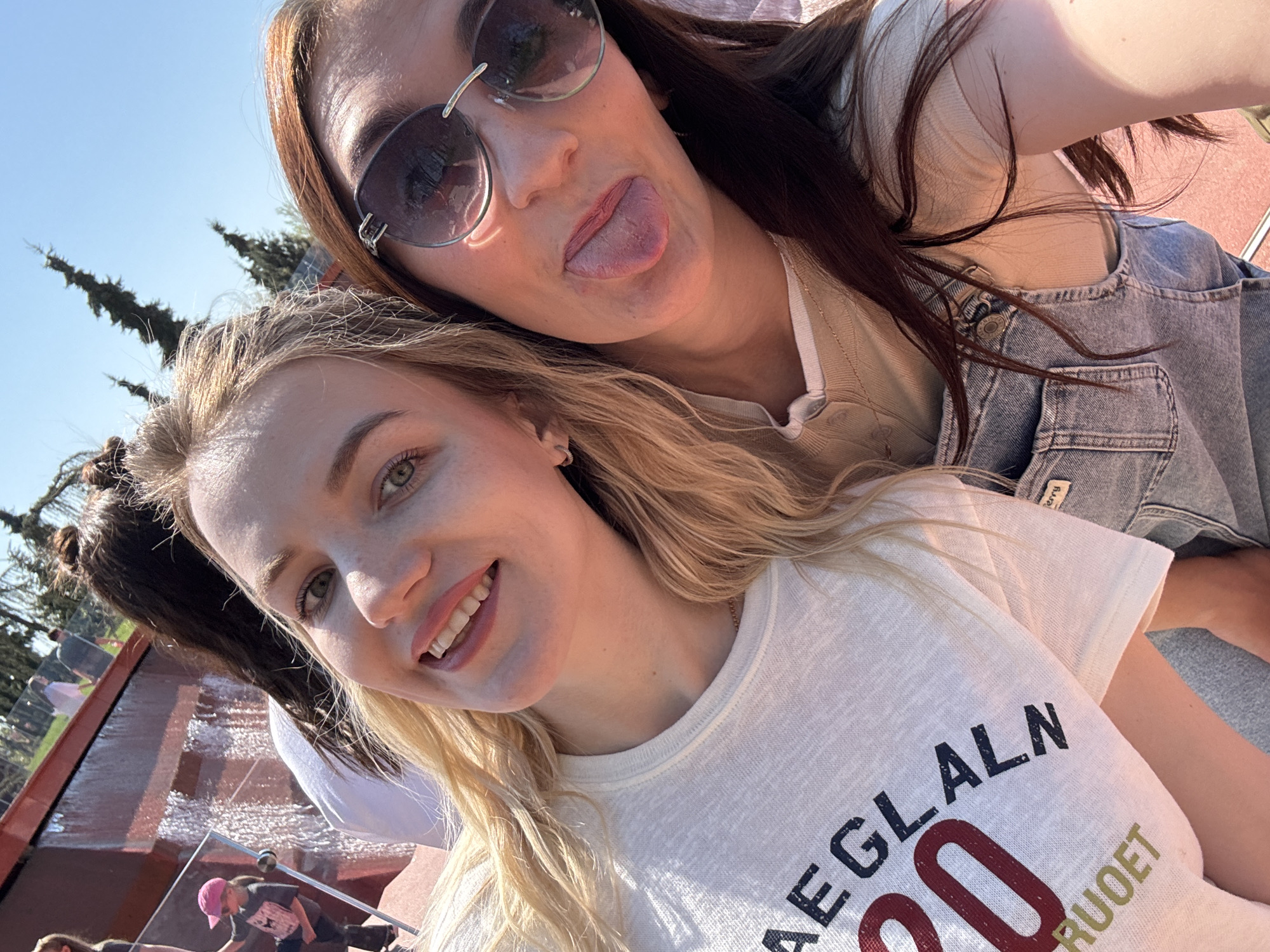 Two women are posing for a selfie. The woman in the foreground has blonde hair and is wearing a white t-shirt with letters on it. The woman in the background has brown hair, is wearing sunglasses, and is sticking her tongue out. They appear to be near a water feature, possibly in a park or public space. The background suggests they may be in Krasnodar, Russia, given the presence of a fountain or waterfall similar to those in the city's parks.