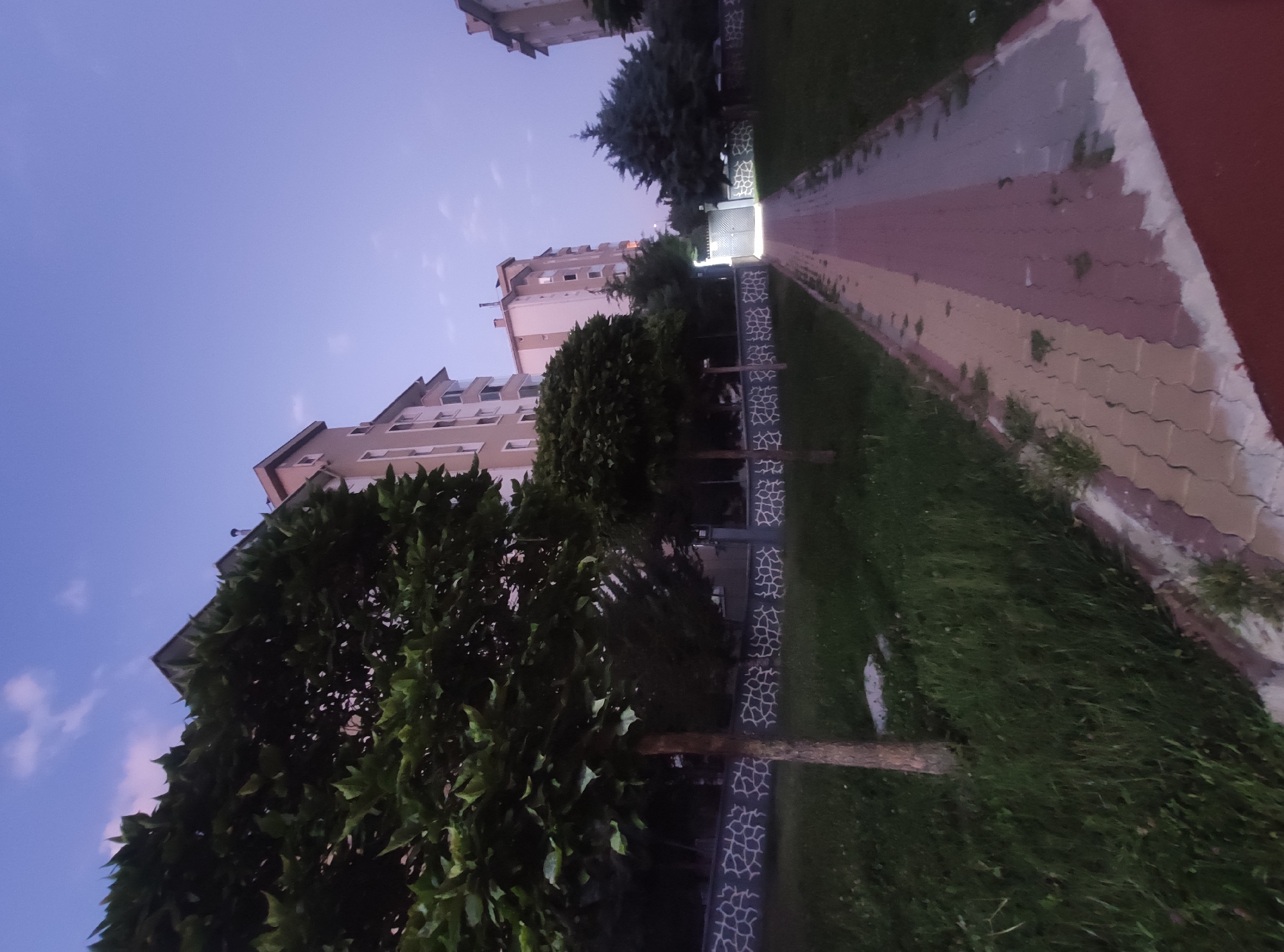 The image depicts a dusk scene in a residential area of Etimesgut, Turkey, likely a courtyard or common area within an apartment complex. In the foreground, a paved walkway with alternating red and beige interlocking bricks leads towards a gate in the background. To the left of the walkway, lush green grass covers the ground, with a slender tree trunk rising from it. Further left, a larger, bushier tree with dark green foliage is prominently featured, its branches casting shadows. Behind the trees, a low decorative wall with a repeating pattern of light grey shapes on a dark background lines the property.
Several apartment buildings dominate the background. The closest building on the left is a multi-story structure with a light beige facade, featuring rows of windows and balconies. To the right of this, another apartment building is visible, taller and with a more prominent section painted light pink or salmon, also with multiple windows. A third, partially obscured apartment building stands to the far right. Above the buildings, the sky transitions from a deep blue near the horizon to a lighter, lavender-grey hue, with faint wisps of clouds. A bright light emanates from the gate in the distance, suggesting it may be open or illuminated. The overall atmosphere is serene and quiet, typical of a residential neighborhood at twilight. There are no people or animals visible in the frame.