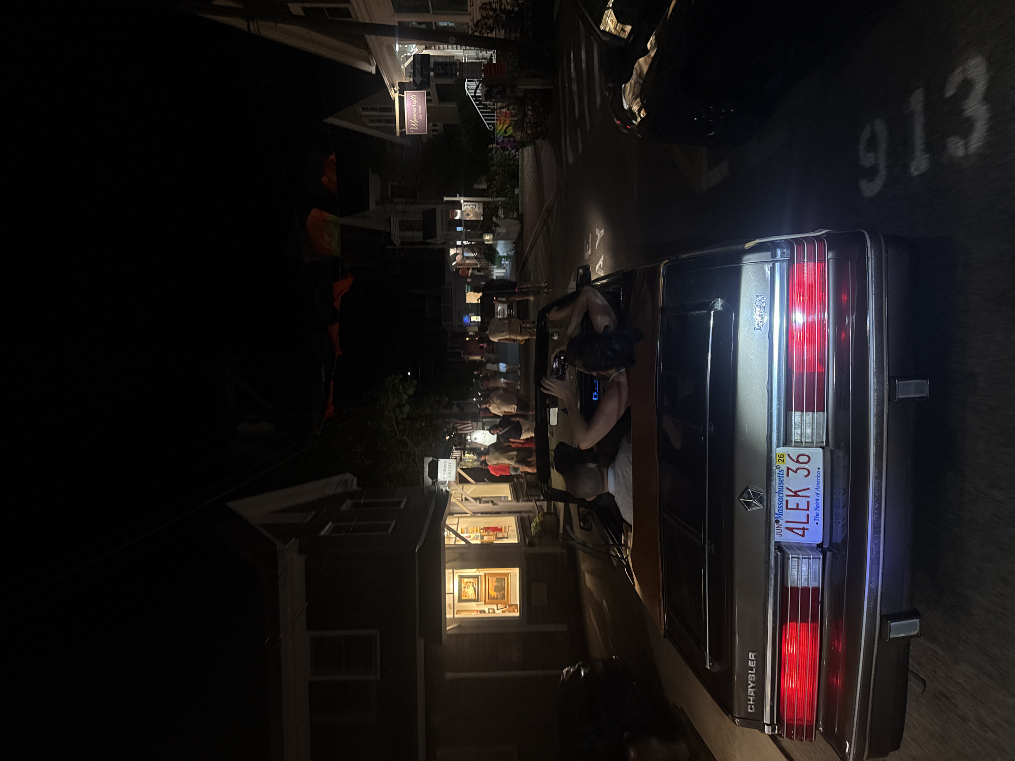 The image shows a nighttime street scene in Provincetown, Massachusetts, USA. In the foreground, the rear of a dark-colored convertible is visible, with a license plate that reads "JUN MASSACHUSETTS 26 4LEK 36 The Spirit of America". Two people are seated in the convertible, with the driver's head and shoulders visible from the back, and a second person, likely female, is leaning back with her head out of the car. The street is lined with buildings, some of which appear to be shops with illuminated windows displaying artwork. People are walking on the sidewalks and standing in front of the businesses. Red and orange triangular flags are strung across the street overhead. The overall atmosphere suggests a lively evening in a commercial district.