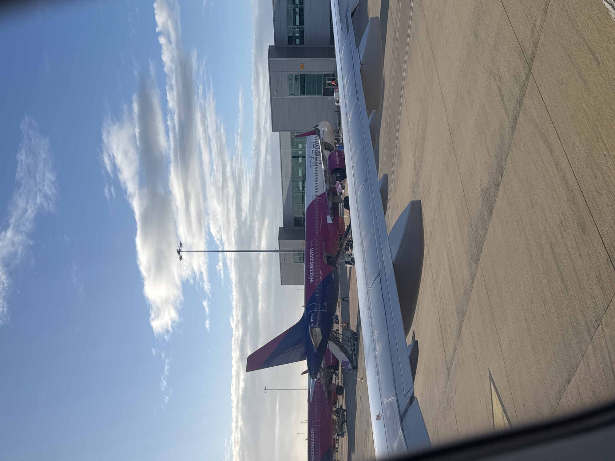 An airport tarmac is shown with several Wizz Air airplanes parked. The foreground features the wing of an airplane, suggesting the photo was taken from a passenger seat. In the background, a modern airport terminal building is visible. The sky is blue with scattered white clouds, indicating fair weather. The ground is paved with concrete, and some yellow markings are visible on it. There are no visible people or specific landmarks to indicate a particular city or location.