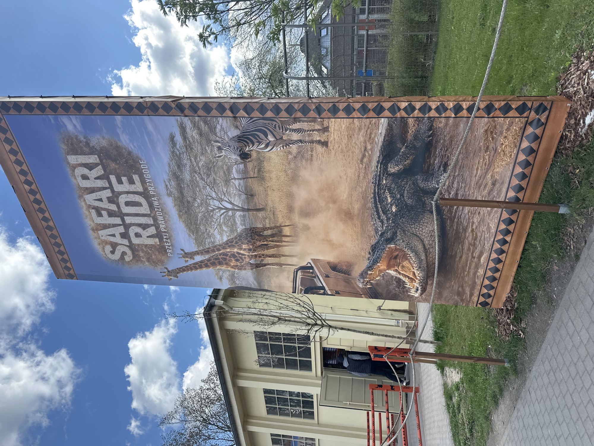 The image shows a large promotional banner for a "Safari Ride – Przeżyj Prawdziwą Przygodę" (Experience a Real Adventure) depicting a safari scene with giraffes, a zebra, a car, and a crocodile. The banner is displayed in front of a light-yellow building with white-framed windows and a doorway, which appears to be a public building due to the presence of a red barricade. The banner’s design includes a border with a repeating diamond pattern. The sky above shows blue with puffy white clouds. The location is likely in Wrocław, Poland, given the Polish language on the banner.
