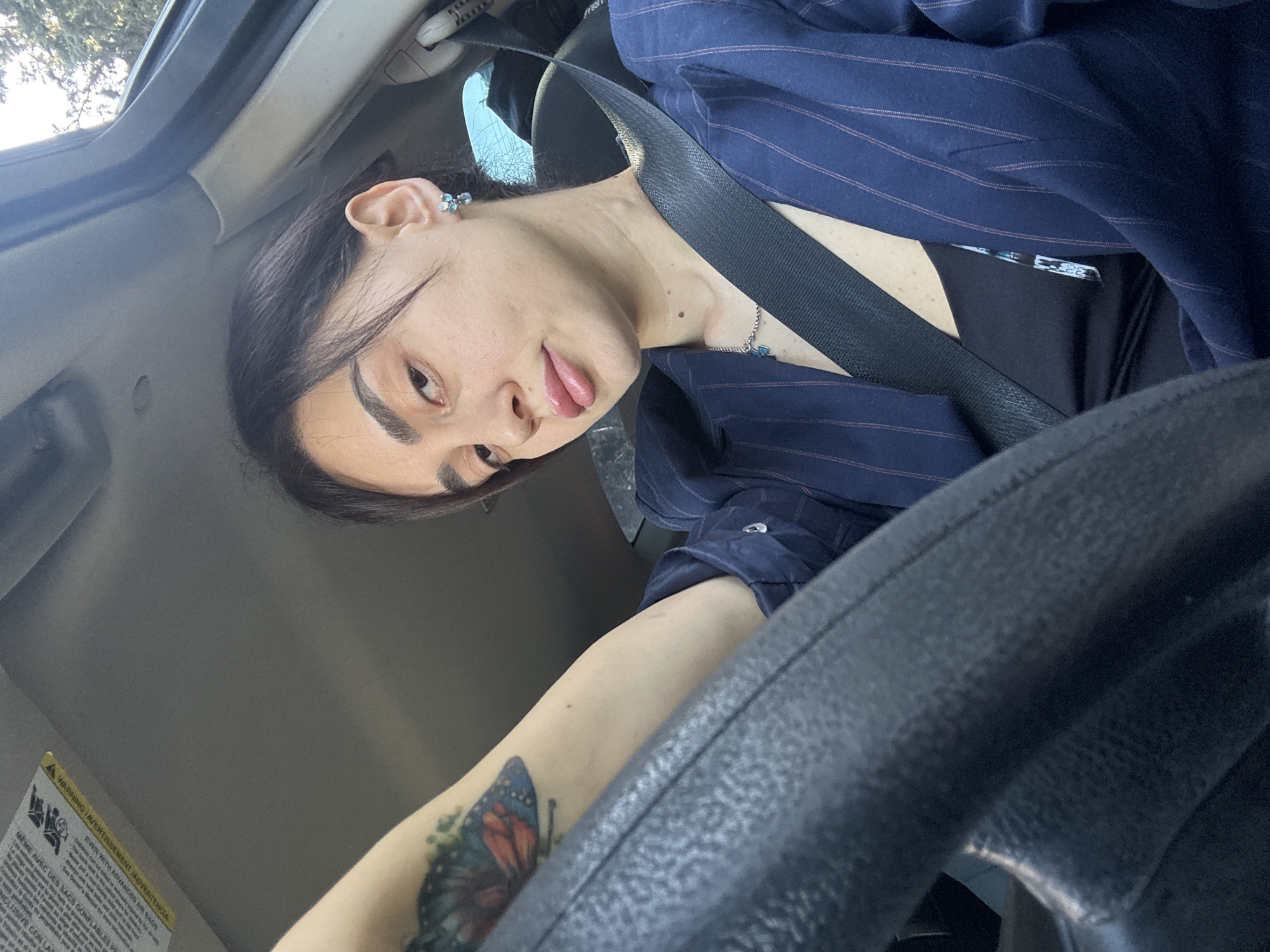 The image shows a person wearing a navy pinstriped blazer and a seatbelt in the driver's seat of a car. The person has dark hair, light skin, and is sticking out their tongue slightly. They are wearing a small earring and a necklace. A tattoo of a butterfly is visible on their arm, which is resting on the steering wheel. Part of the car's interior, including the dashboard and the sun visor with warning labels, are also visible. The background through the windshield shows trees. It's impossible to tell if the image was taken in Tbilisi, Georgia without additional information.