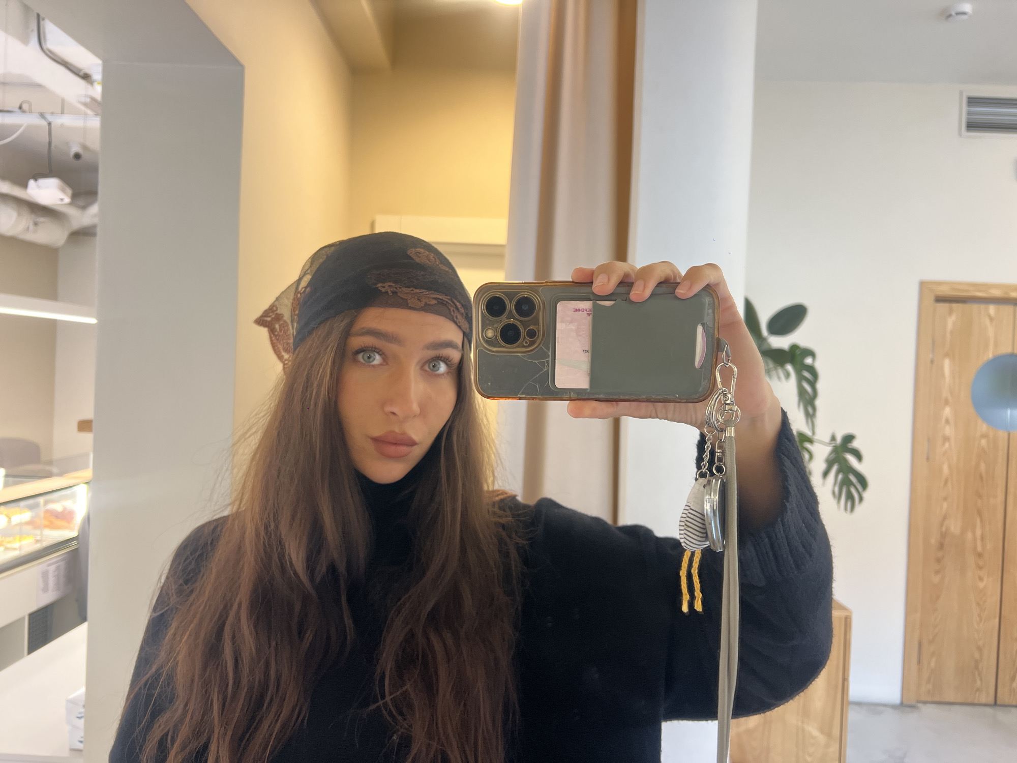 The image shows a woman taking a selfie in a mirror in a building, likely in Vladivostok, Russia, though this cannot be confirmed with certainty based solely on the image. The woman has long brown hair and is wearing a dark-colored turtleneck sweater and a dark scarf or head covering. She is holding a smartphone in her hand, and the phone case appears to be cracked. There is a card inserted in the phone case. Attached to the phone is a keychain with various small objects. The background reveals a brightly lit interior space. To the left, there is a glass display case with baked goods. In the mirror's reflection, there's a plant, a door, and an air conditioning vent. The overall environment appears to be modern and well-maintained.