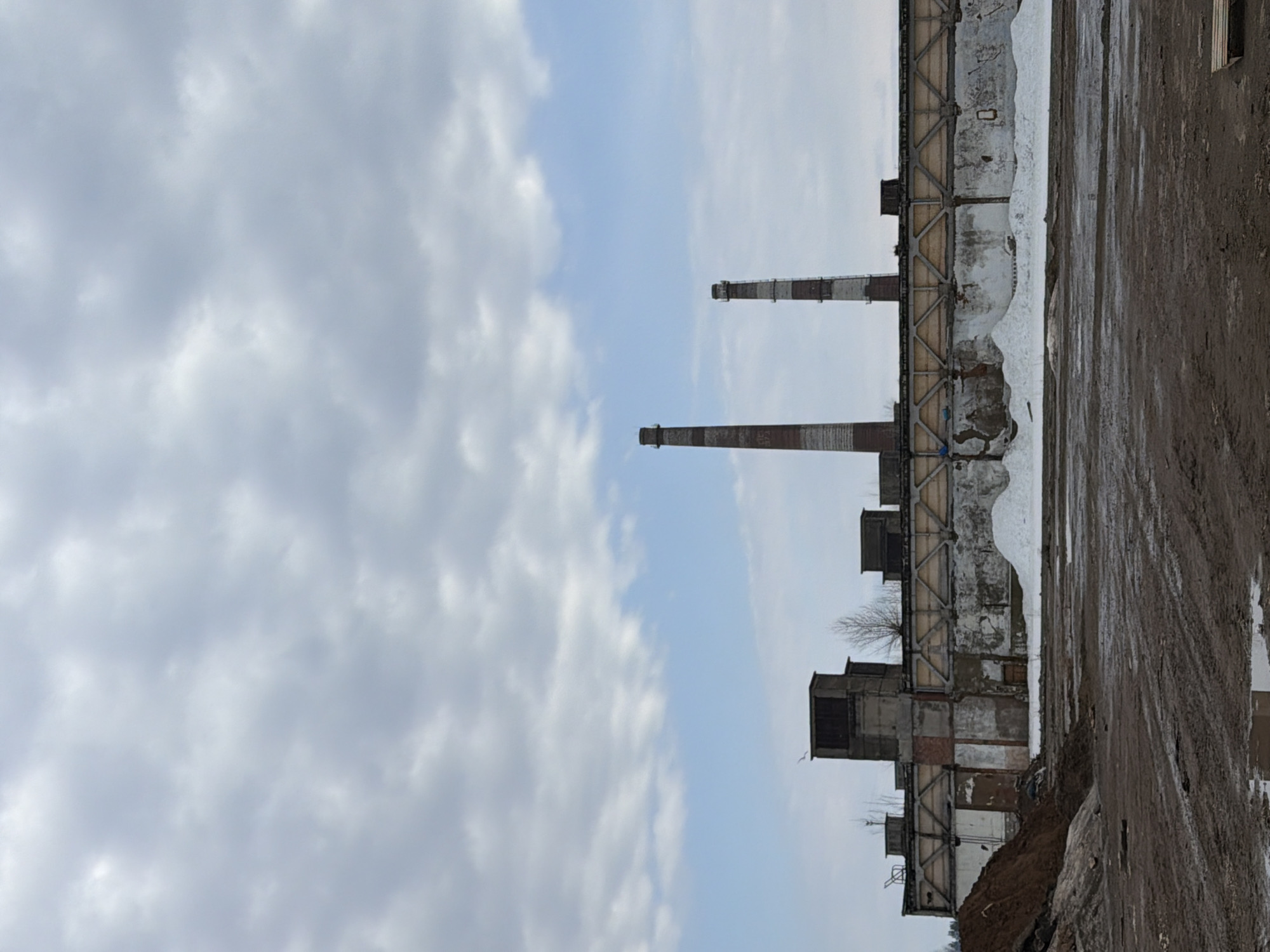 The image shows an industrial landscape in what appears to be the city of Chaikovsky, Russia. The foreground features a muddy ground. The right side shows a tall, decaying industrial building. Behind this building are two prominent smokestacks. The sky is overcast with a mix of white and gray clouds.