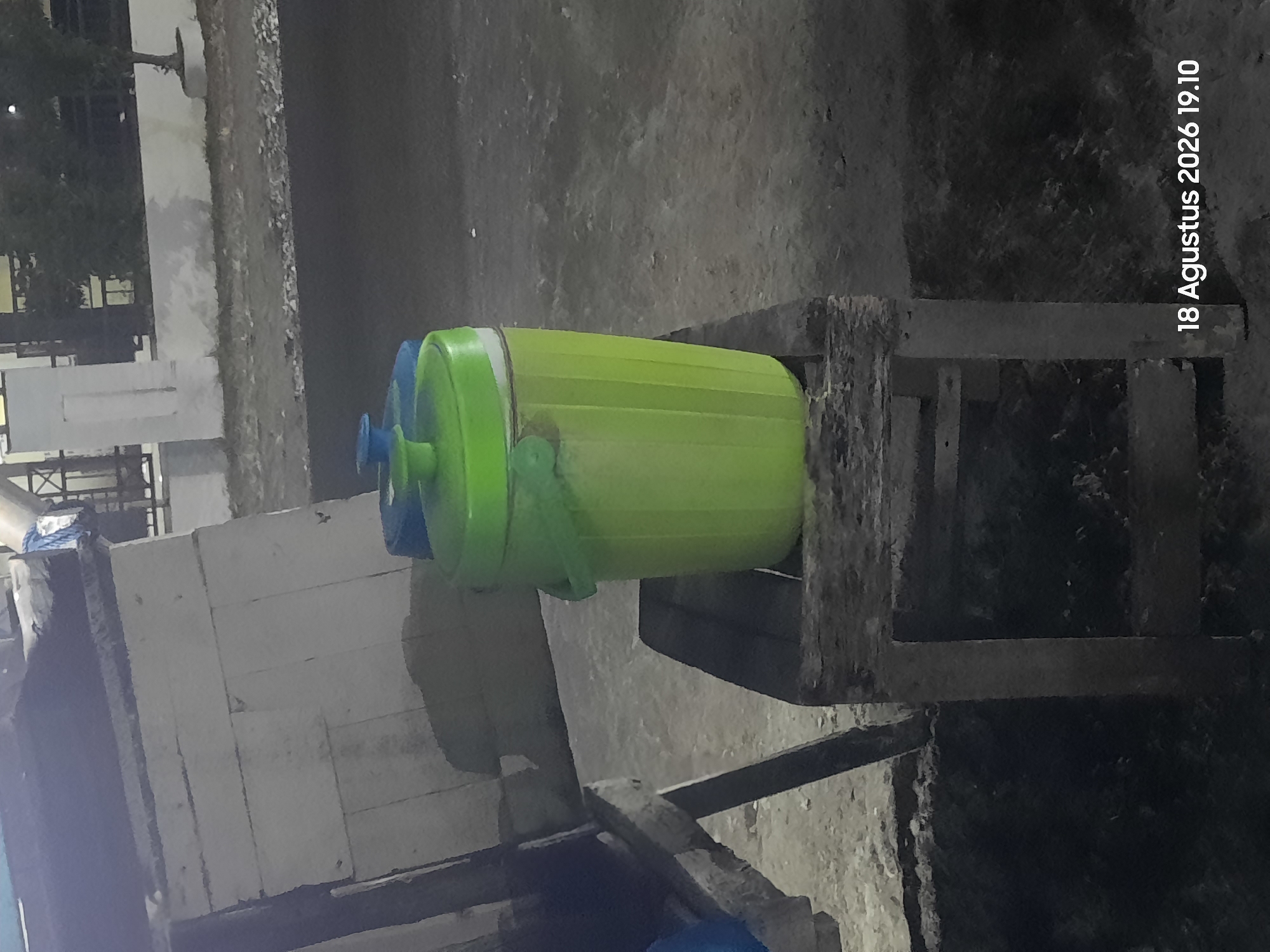 In Sukokerto, Indonesia, on August 18, 2026, at 19:10, a nighttime scene unfolds featuring two large water dispensers. The primary subject is a bright green, ribbed water dispenser with a green lid and handle, placed centrally on a rustic wooden stool. To its left, a portion of a blue water dispenser is visible, also with a lid. The dispensers are positioned outdoors on a rough, uneven ground surface that appears to be paved or a dirt path. In the background, a white building with visible brickwork and architectural features suggests a residential or commercial structure. Trees and foliage are dimly lit, indicating an outdoor setting at dusk or after dark. The overall atmosphere is subdued, with artificial lighting casting shadows and highlighting the textures of the objects and environment. The wooden stool appears weathered and is made of rough-hewn planks.