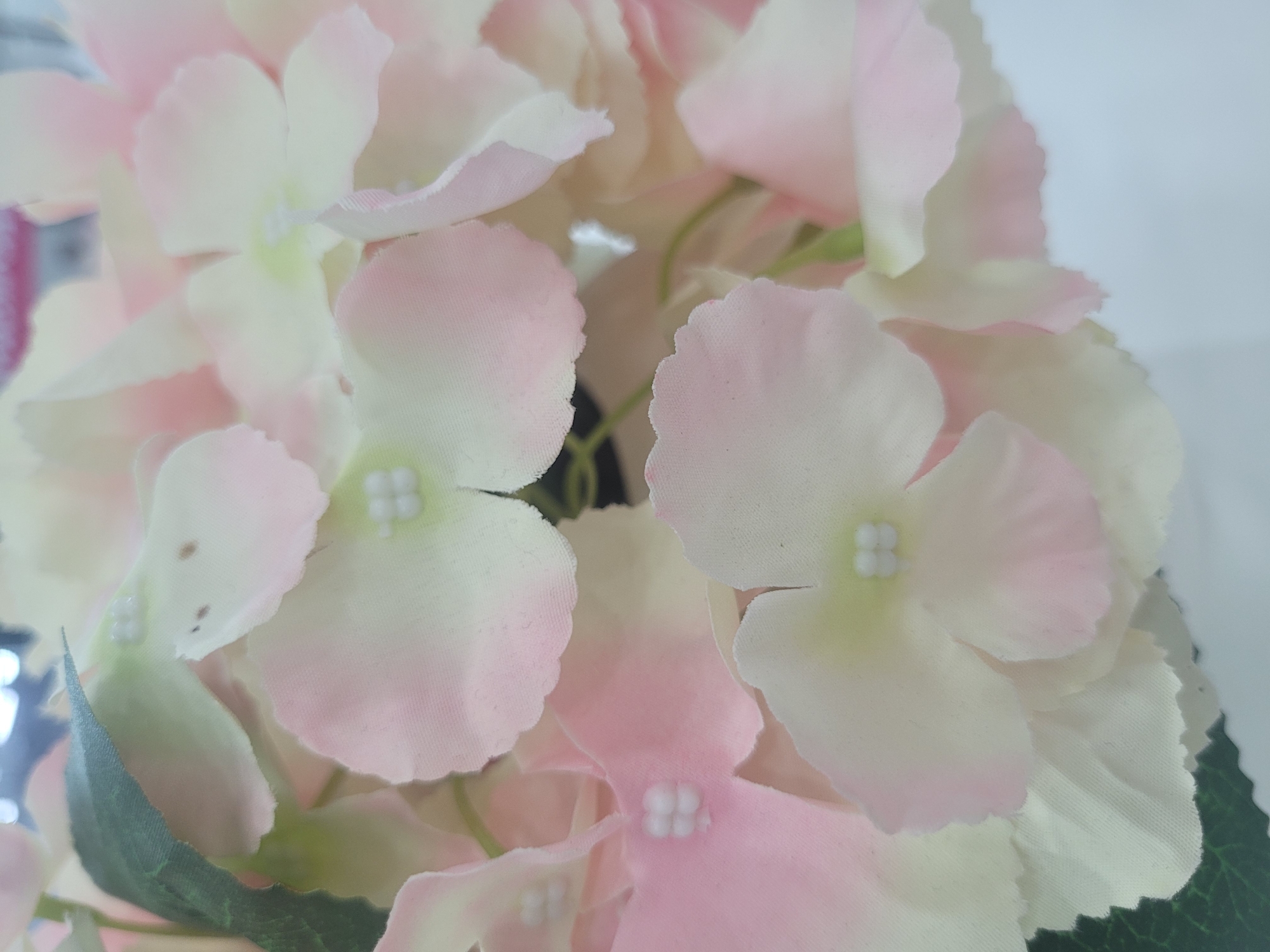 A close-up, overhead shot showcases a cluster of artificial hydrangeas. The flowers are a delicate blend of pale pink and white, with soft, rounded petals. Each flower head features a cluster of tiny white beads at its center, mimicking stamen. A few green leaves are visible, one prominent dark green leaf in the lower left corner, with another hint of green peeking from behind a flower. The background is a plain, light-colored surface, possibly a wall or table, with a slightly out-of-focus dark element visible in the upper center. The lighting appears bright and even, suggesting an indoor setting. No text is visible in the image. The focus is entirely on the textural and color details of the artificial flowers.