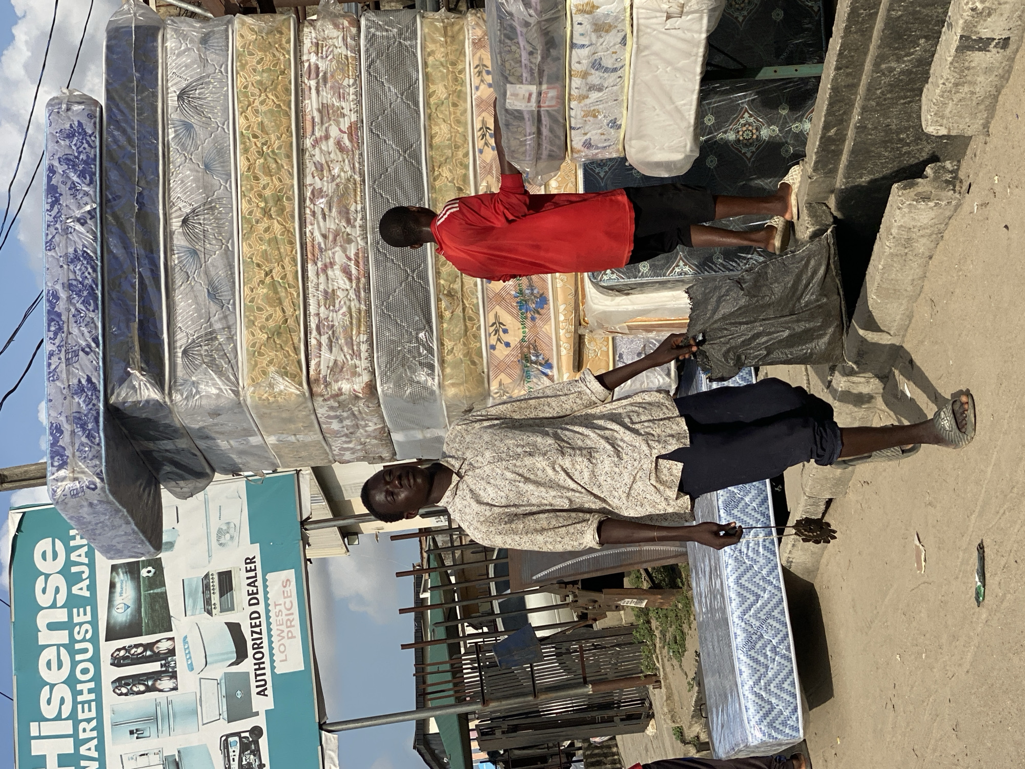 The image shows a street scene in Ado, Nigeria, with a Hisense warehouse billboard on the left. Two men are present, one wearing a red shirt and the other a patterned shirt with dark pants. They appear to be involved in selling or transporting mattresses. A stack of mattresses wrapped in plastic is visible in the background. The ground is bare and the sky is partially visible.