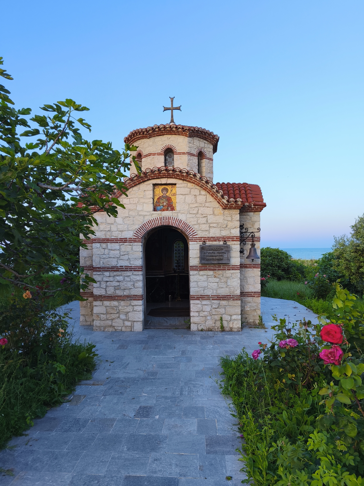 The image shows a small, stone chapel with a red-tiled roof, topped by a cross. The chapel is adorned with a mosaic above the arched entrance and a bell mounted on the side. A paved path leads to the chapel, flanked by lush green bushes and vibrant pink and red roses. In the background, the calm blue sea stretches towards the horizon under a clear sky. This is the Chapel of Saint Paraskeva in Tsarevo, Bulgaria, situated close to the Black Sea coast.
