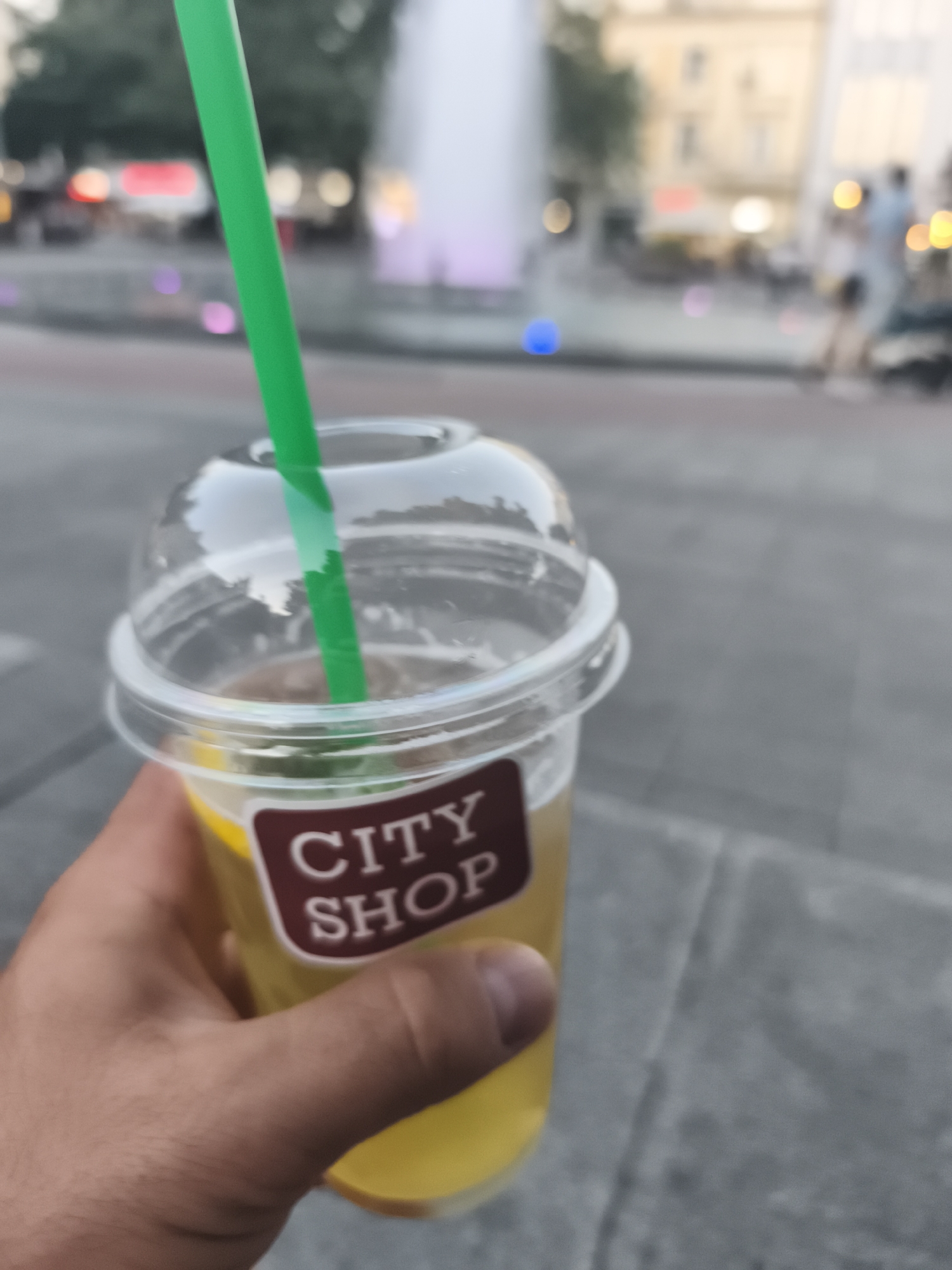 A close-up, shallow depth-of-field shot shows a hand holding a clear plastic cup filled with a yellow beverage. A green straw protrudes from the top of the cup, which is covered with a clear plastic lid. A brown label on the cup reads "CITY SHOP". The background is blurred but suggests an outdoor urban setting with a fountain and buildings. This scene was captured in Plovdiv, Bulgaria, likely in the pedestrian zone near the Roman Stadium.