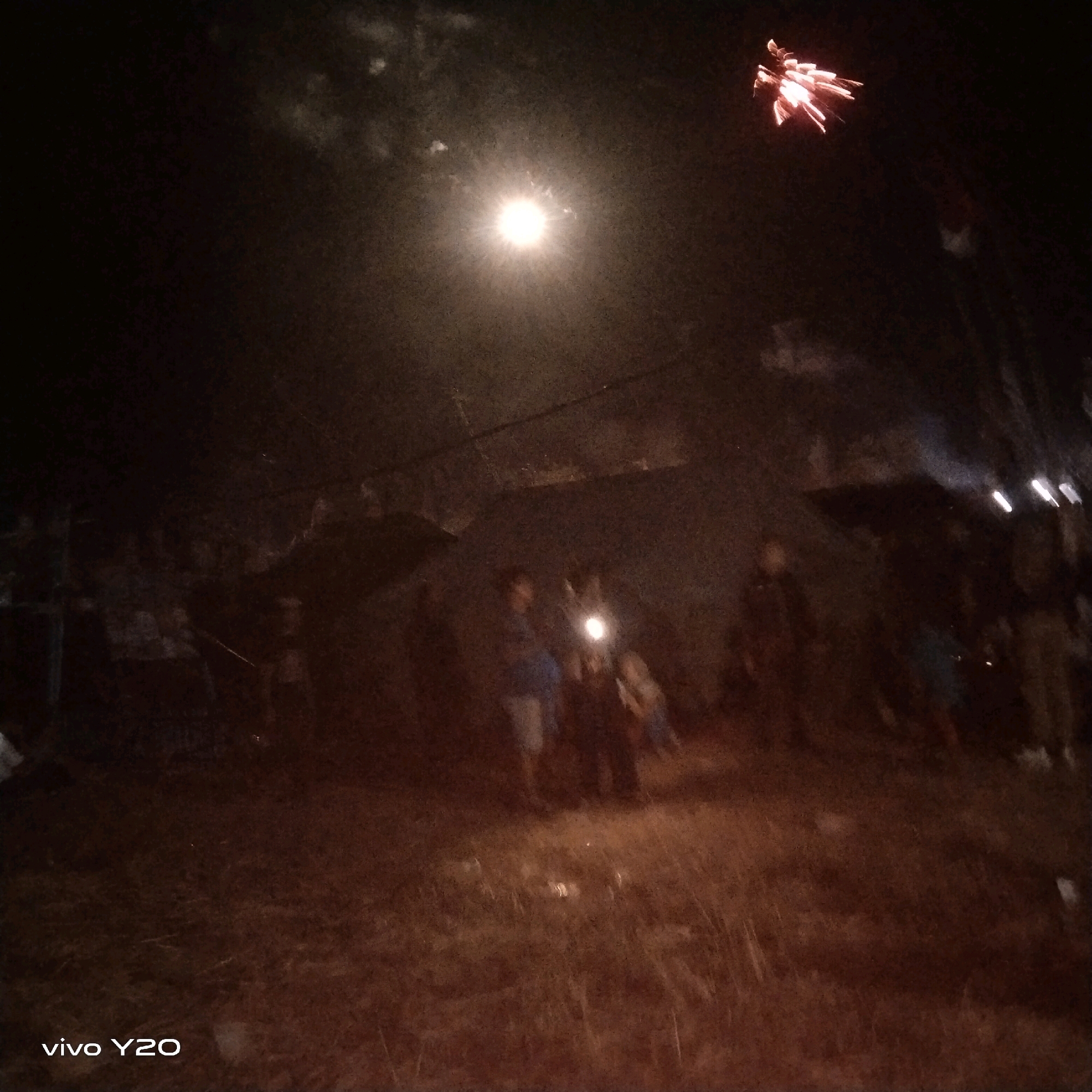 A blurry, low-light photograph captures a night scene with a bright light source illuminating the center, possibly a streetlight or a camera flash. In the upper right corner, a small, bright object streaks across the dark sky, resembling a firework. Below the bright light, several indistinct figures are visible, some appearing to be standing and watching. The ground appears to be rough and uneven, possibly dirt or grass. A watermark in the lower left corner reads "vivo Y20". The overall impression is of a gathering or event taking place at night, with some form of illumination and possibly fireworks.