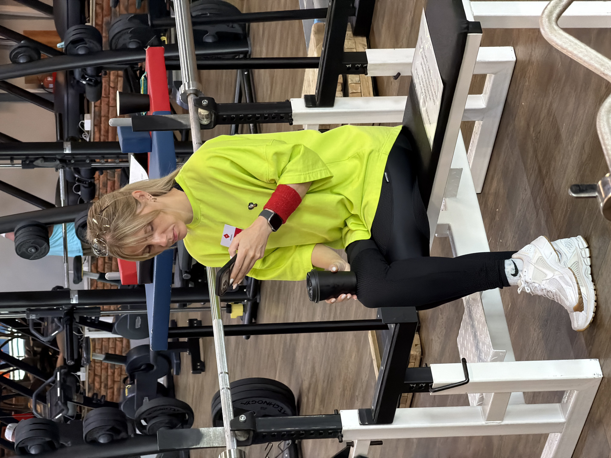A Caucasian woman with blonde hair is sitting on a weight bench in a gym. She is wearing a bright green long-sleeved shirt, black leggings, and white athletic shoes. She is holding a black smartphone in her right hand and a black paper cup in her left hand. A red wristband is visible on her left arm. The gym appears to be equipped with various weight machines and equipment. The floor is made of wood-look laminate. The woman is looking at her phone. There is a metal barbell across the bench in front of her. The background shows gym equipment and brick walls. This scene is likely located in the city of Chaikovsky, Russia.