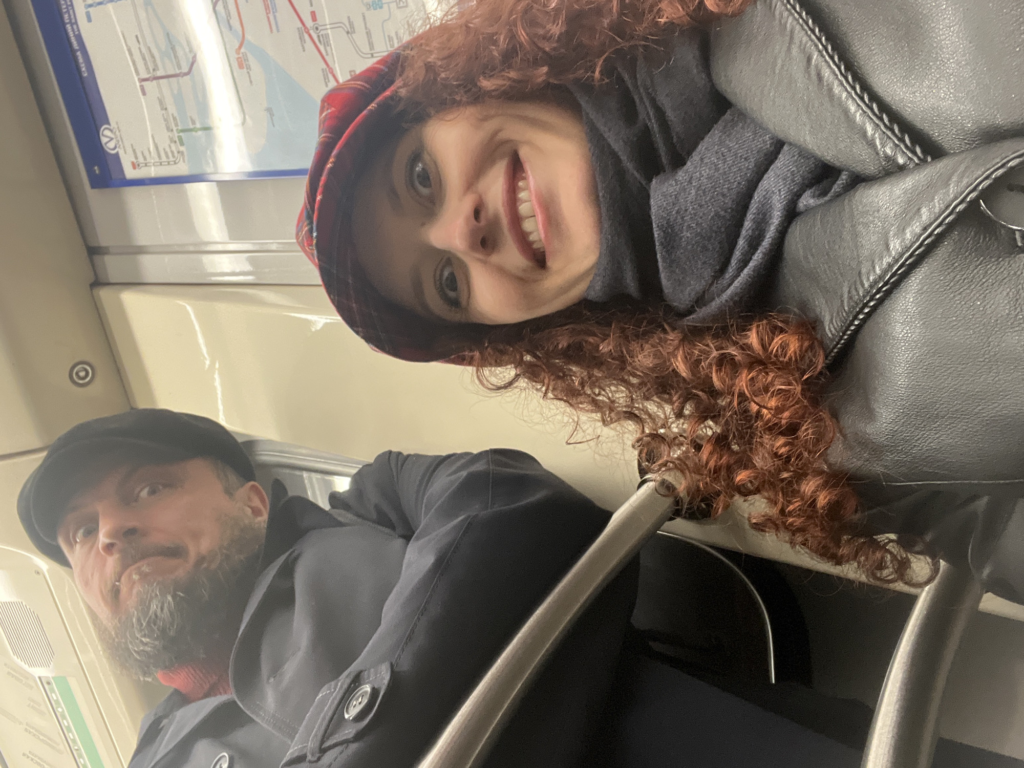A photo of a man and a woman in what appears to be a subway car in Saint Petersburg, Russia, based on the design and signage. The woman has curly auburn hair and is wearing a red plaid hat, gray scarf, and a black leather jacket. She is looking at the camera and smiling broadly. The man is wearing a black cap and a black coat and has a gray beard. He is looking up and slightly to the left. The background shows a map of the Saint Petersburg metro system and the interior of the subway car, including the seats, handrails, and wall panels.