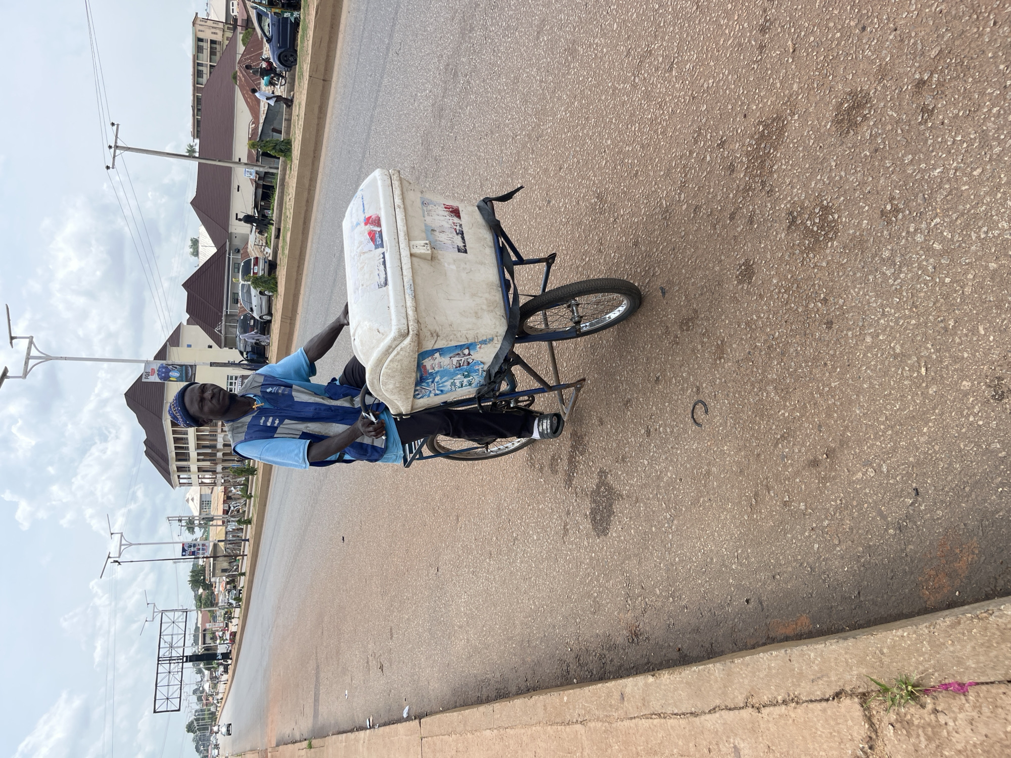 A man is riding a tricycle with a large white cooler attached to the back. He is wearing a blue shirt and vest and a blue cap. The tricycle is on a paved road in an urban area. Buildings and power lines are visible in the background. The sky is cloudy. The man appears to be looking towards the camera. The time of day is likely daytime. The location is Bukuru, Nigeria. There is some text visible on the cooler, but it is difficult to read.