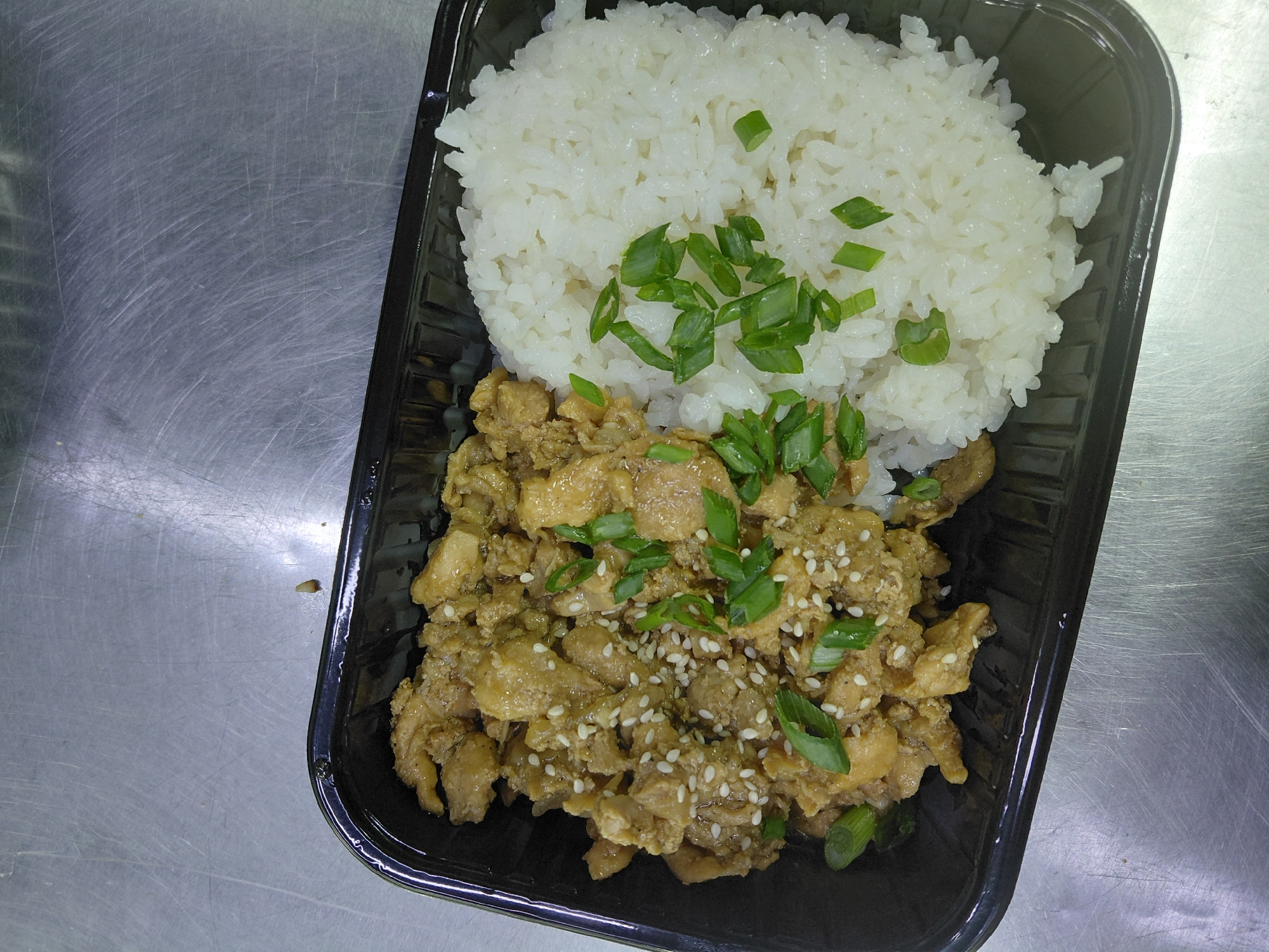 A black plastic food container holds a meal on a scratched metallic surface. The container is divided into two compartments. The left compartment contains a serving of stir-fried chicken pieces seasoned with sesame seeds and garnished with chopped green onions. The chicken appears to be coated in a light brown sauce. The right compartment contains a mound of white rice, also garnished with chopped green onions. The overall lighting is overhead and slightly diffused, creating some glare on the metallic surface. No people are visible in the image. The location is identified as Aktau, Kazakhstan, based on information associated with the image.