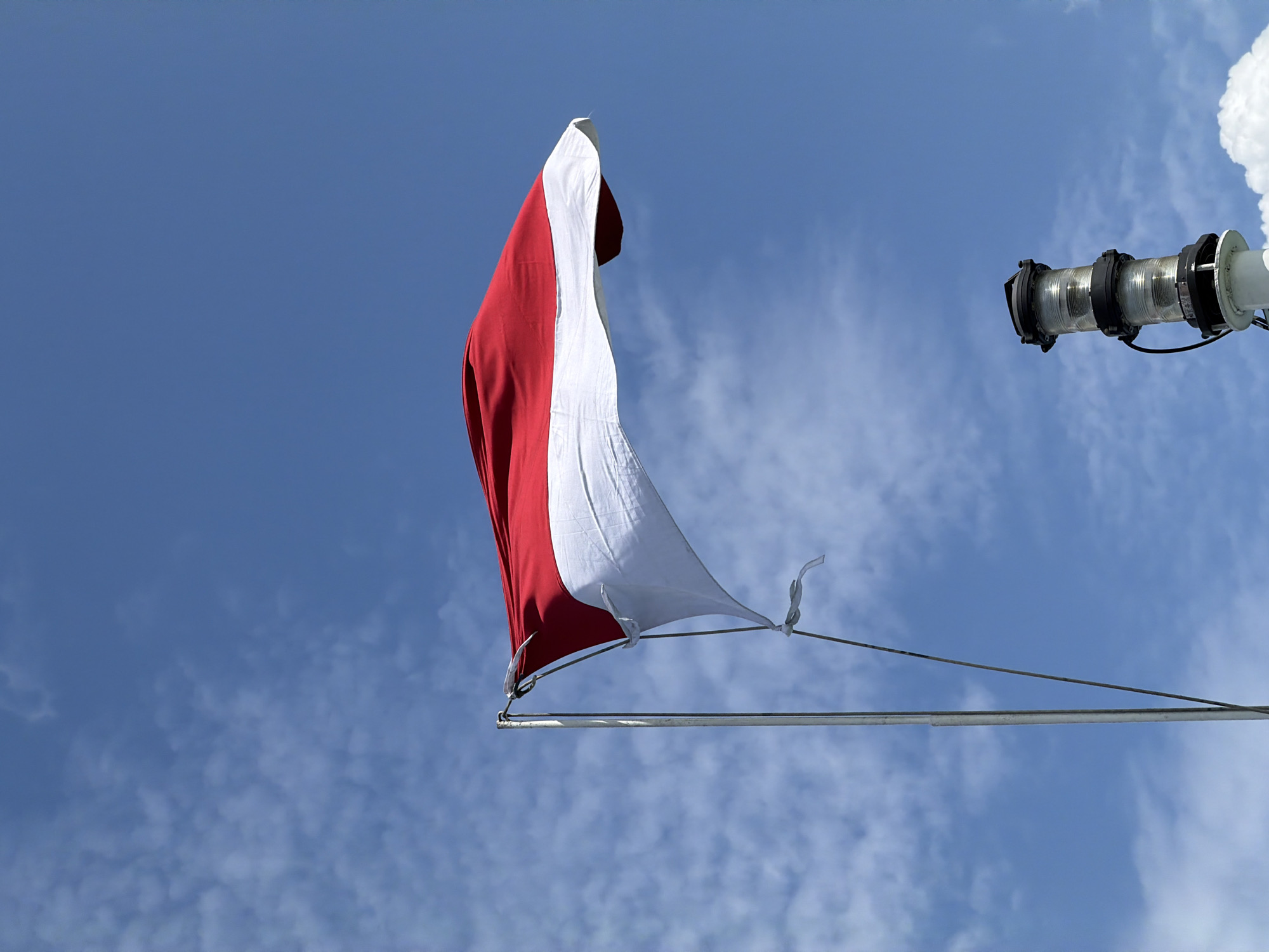 The image shows the Indonesian flag waving in the wind against a bright blue sky with scattered white clouds. The flag, which is red and white, is attached to a metal flagpole. To the lower right, a ship's navigation light is visible. The image appears to be taken from a low angle, looking up at the sky. The scene is likely located in or near the harbor of Ketapang, Indonesia, a coastal city known for its maritime activities. There are no people visible in the image.