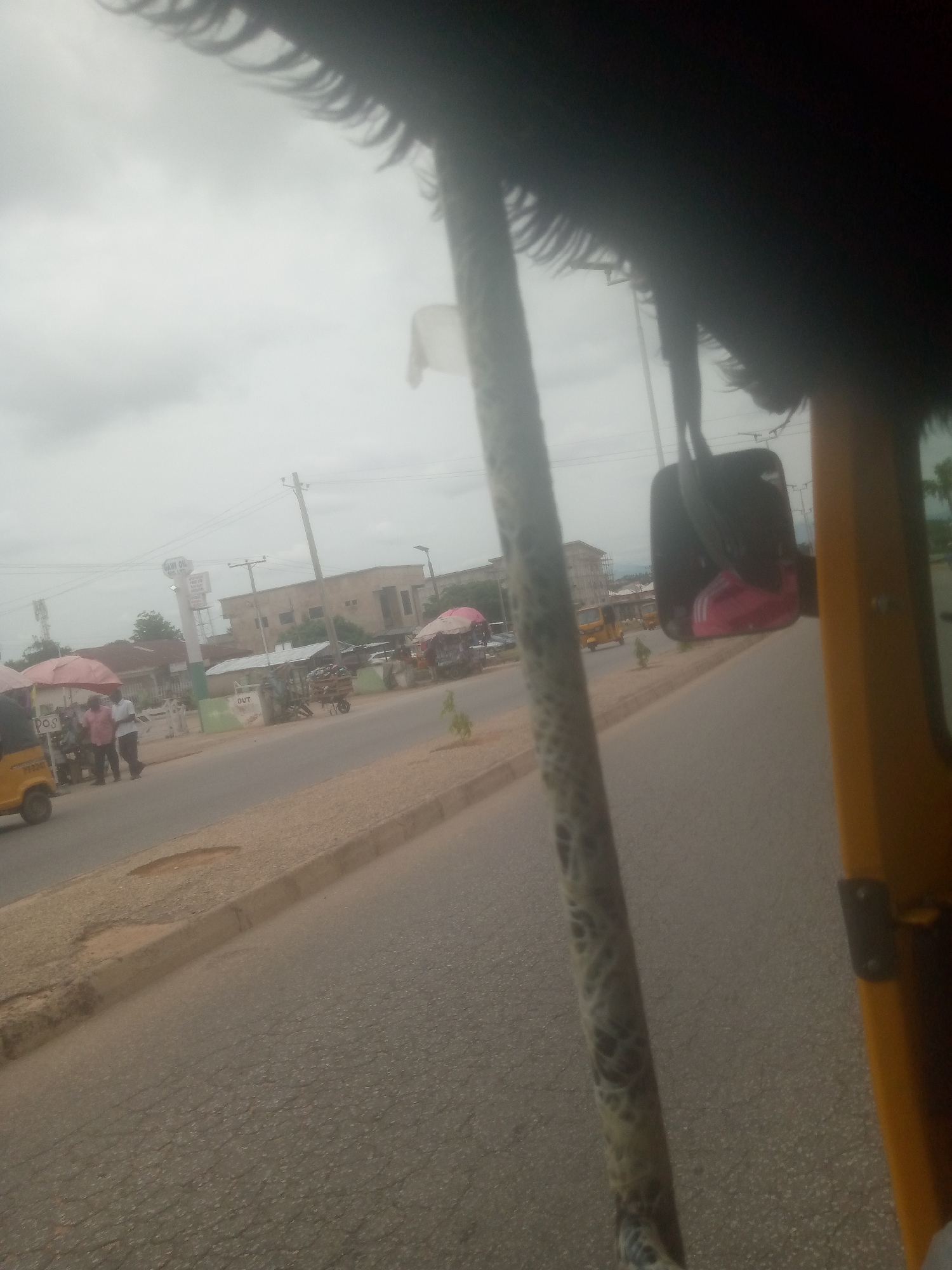 The image depicts a street scene in Jalingo, Nigeria. The viewpoint appears to be from inside a three-wheeled vehicle, likely a motorized rickshaw, as indicated by the yellow paneling and the interior structure visible in the foreground. Outside, a paved road runs alongside a sidewalk with a curb.
On the sidewalk, several individuals are gathered around stalls, some covered by umbrellas. One stall has a "POS" sign. There are also carts with goods displayed for sale. In the background, buildings of various sizes and designs are visible, along with utility poles and electrical wires. A tall sign, possibly for a business, with the text "SAWI OIL" and "ENTERPRISE" is prominent.
The sky is overcast and appears to be a pale gray, suggesting it is either early morning, late afternoon, or a cloudy day. The overall impression is of a typical street scene in a bustling town or city, with commerce and daily life unfolding on the roadside. The lighting is diffused, and there are no strong shadows, consistent with a cloudy sky.