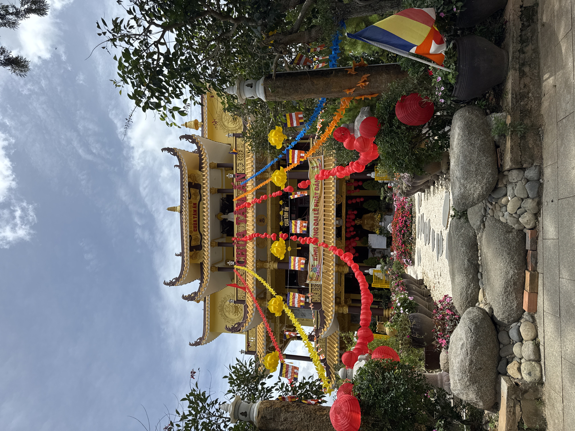 This is an exterior, eye-level, medium shot of a Buddhist temple in Da Lat, Vietnam, under a partly cloudy sky. The temple, a multi-tiered structure painted in yellow and gold, is adorned with vibrant red and yellow paper lanterns and prayer flags. In the foreground, a stone path leads towards the temple, flanked by large rocks, potted plants, and blooming flowers. A large Buddhist flag, featuring five horizontal stripes in blue, yellow, red, white, and orange, is visible on the right side of the image, planted in a large black pot. The scene conveys a festive and spiritual atmosphere, likely during a holiday or celebration.