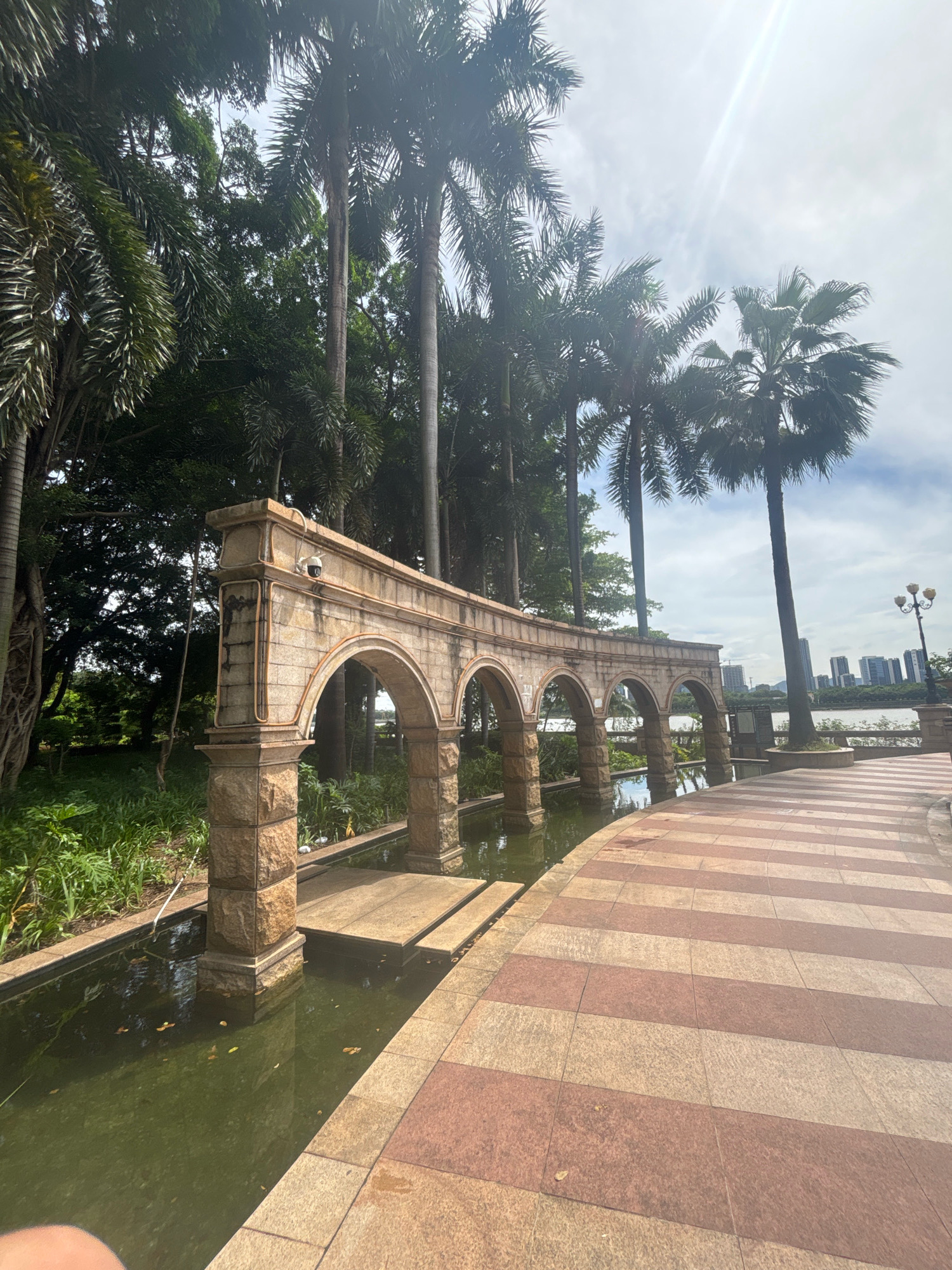 The image depicts a stone arcade with multiple arches, set against a backdrop of palm trees and lush greenery. The arcade appears to be situated in an outdoor recreational area, possibly a park or waterfront promenade, in Nanhai District, China. A walkway with alternating red and beige paving stones curves around the arcade, bordering a shallow body of water. In the distance, a city skyline with modern buildings is visible across a body of water. The sky is partly cloudy, with the sun casting a glare on the scene. There are no people visible in the image, and the overall atmosphere is serene and natural. A security camera is mounted on the top section of the arcade.