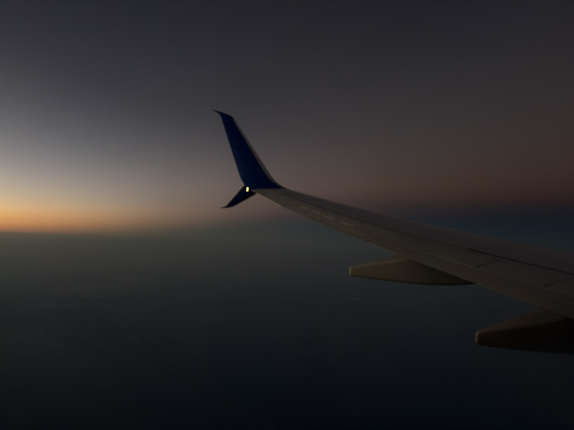 A view from an airplane window shows the plane's wing against a backdrop of the earth and sky at dusk or dawn. The sky is transitioning from a dark blue/purple at the top to a lighter orange hue near the horizon. The earth below is dark and indistinct. The plane's wing is predominantly white/grey, with a blue tip. A small light is visible on the wing tip. The location and details in the city of Port Fourchon, United States, cannot be determined from the image.
