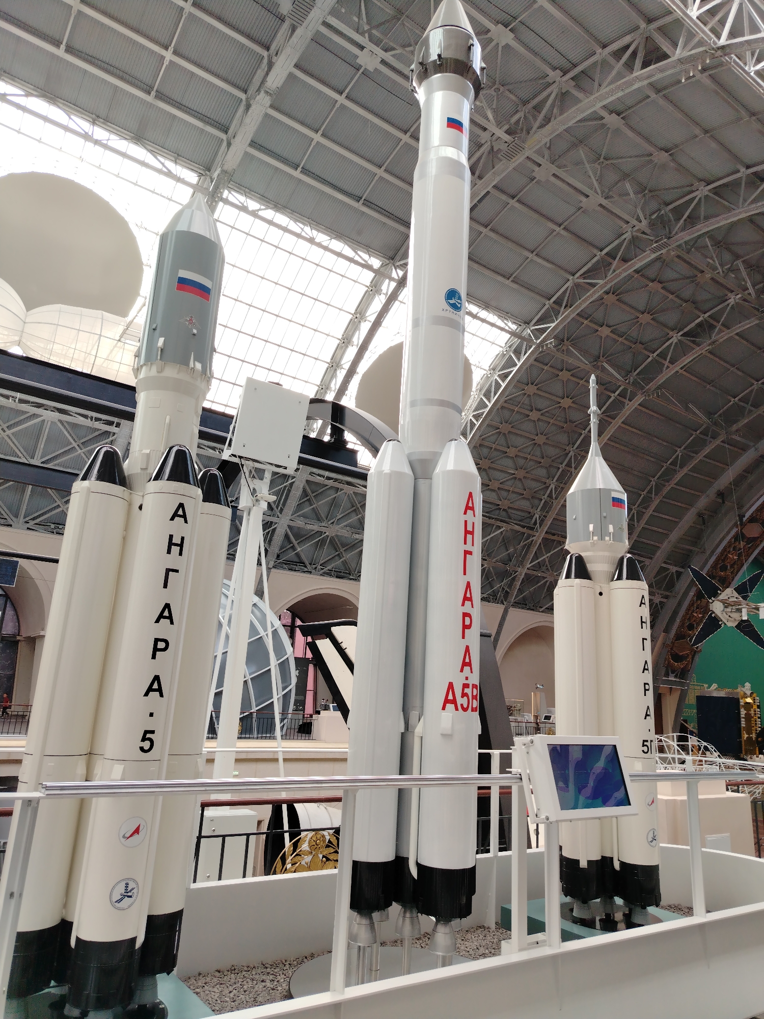 This image depicts models of the Angara rocket family displayed indoors, likely in a museum. Several rockets are visible, with the most prominent being a large white rocket in the center, identified by Cyrillic lettering on its side. To the left is a smaller rocket with a gray upper stage and a Russian flag emblem. To the right, another smaller rocket with a similar design is partially visible, also featuring the Russian flag. The rockets are mounted on stands, and a railing runs in front of them. Behind the rockets, a large, vaulted ceiling with exposed metal beams and translucent panels can be seen, suggesting a spacious exhibition hall. The overall environment appears to be a museum or exhibition space dedicated to space exploration. The image does not contain any people.