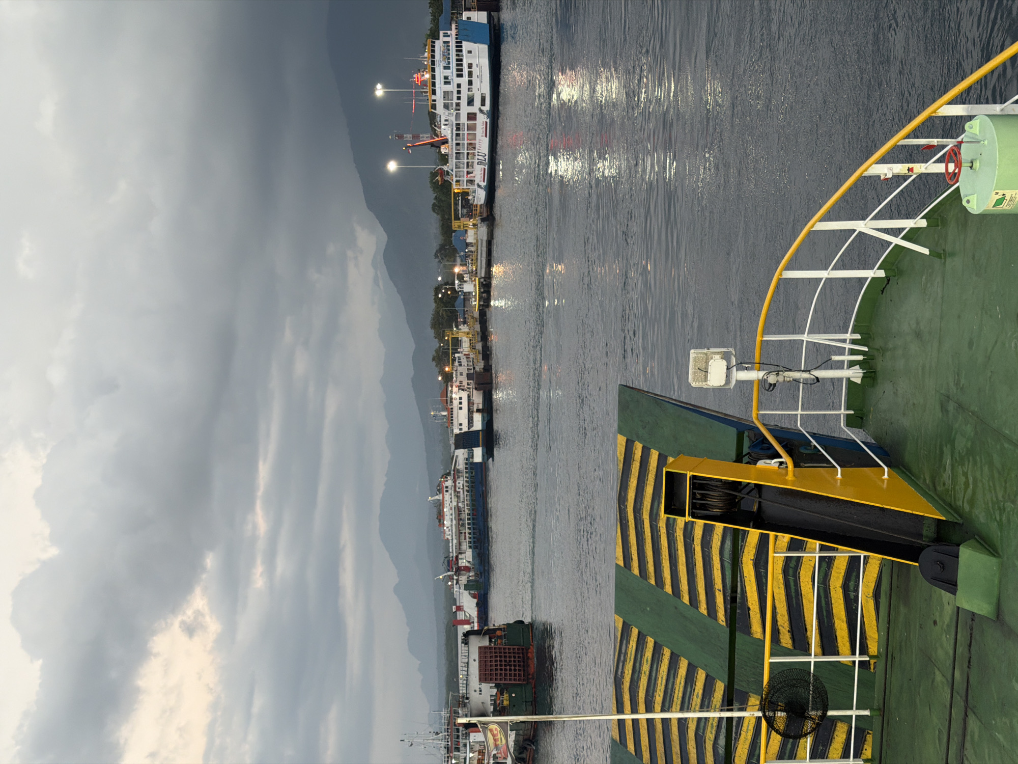 The image depicts a ferry terminal in Melaya, Indonesia, at dusk or dawn. In the foreground, the deck of a ferry is visible, featuring a ramp with yellow and green chevron markings, a black winch, and a green cylindrical object with a red wheel. White railings with a yellow handrail line the edge of the deck. In the middle ground, several ferries are docked at a pier. One prominent white ferry, branded "RLU," is brightly lit. Other ferries, some appearing older and industrial, are also present. The background shows a shoreline with distant mountains under a cloudy, overcast sky. The water is calm, reflecting the lights from the terminal and ferries. The scene suggests a busy port environment, likely a transportation hub for vehicles and passengers.