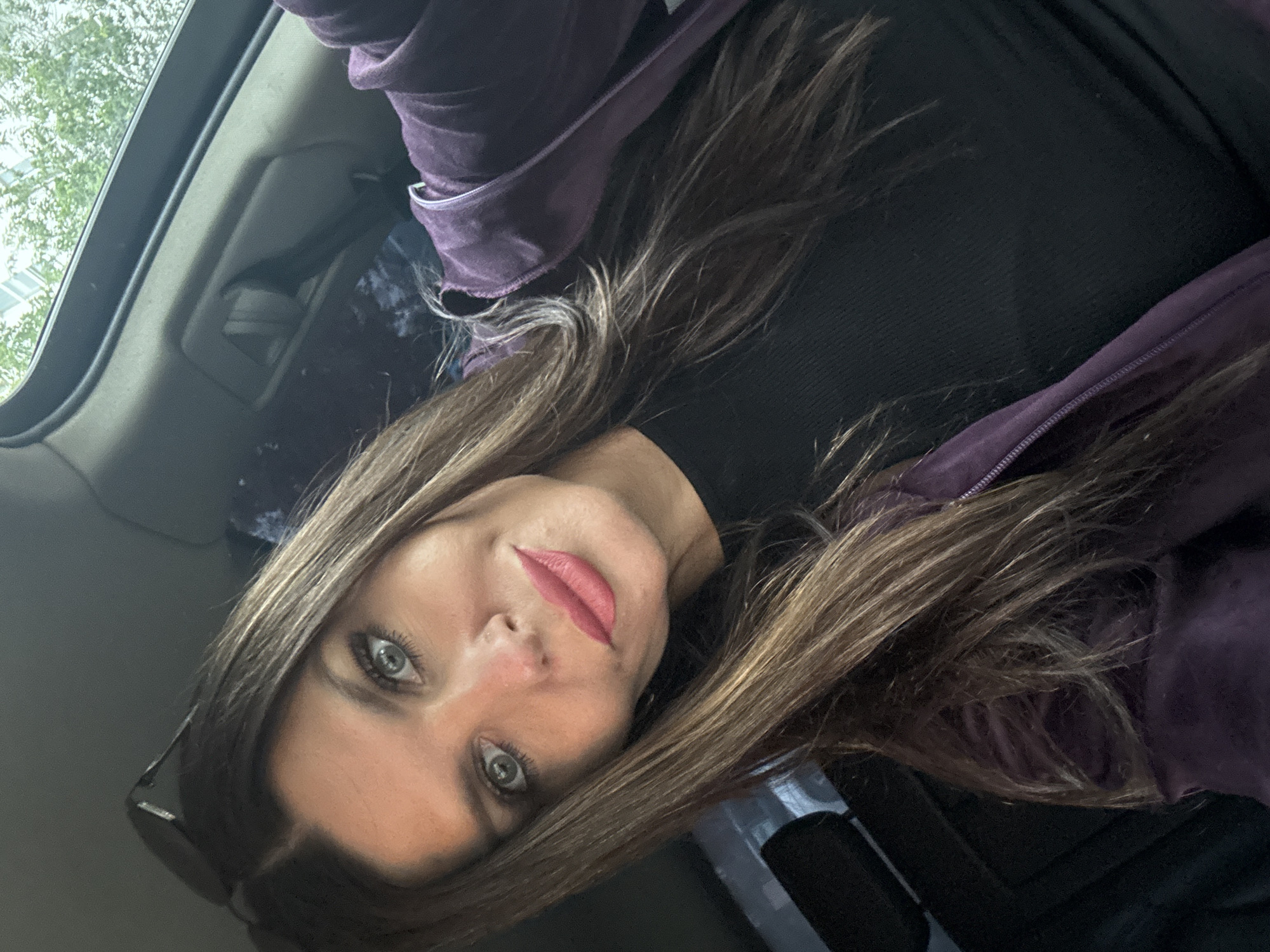The media file shows a close-up, upside-down selfie of a woman. She has long, brown hair with lighter streaks and is wearing a black top and a purple jacket. Her makeup includes pink lipstick and mascara. She appears to be inside a vehicle, as suggested by the interior elements visible, such as a sunroof and part of the car's interior paneling. There are no visible text elements in the image. The lighting suggests it is daytime.