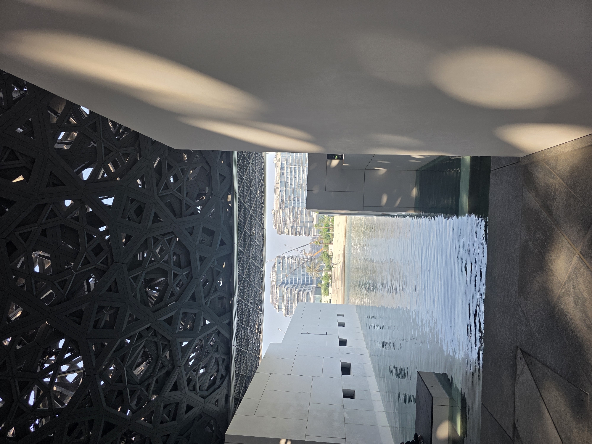 The image depicts the Louvre Abu Dhabi, a universal museum located on the Saadiyat Island in Abu Dhabi, United Arab Emirates. The scene is captured from a low angle, looking through a water feature towards the museum's iconic dome and the surrounding cityscape.
The foreground shows a body of water, likely part of the museum's architectural design, with ripples on its surface reflecting the light. To the left, a white wall with several square openings is visible, extending into the water. A dark, rectangular structure, possibly a platform or step, is partially submerged in the water to the far left. The ground in the immediate foreground is paved with large, grey tiles, some of which are illuminated by dappled sunlight, creating geometric shadows.
Above the water and walls, the intricate latticework of the Louvre Abu Dhabi's dome is prominently featured. This complex geometric structure, made of dark metal, allows sunlight to filter through, creating patterns of light and shadow. To the right, a large, smooth, light-colored wall dominates the frame, with broad, diagonal shadows cast by the dome or an external structure.
In the background, across the water, modern buildings rise, some appearing to be under construction, indicated by visible cranes. Palm trees and other greenery are also present, suggesting an outdoor setting. The sky is a pale blue, indicating daylight. The overall impression is one of modern architecture integrated with natural elements, bathed in the warm light of the afternoon sun.