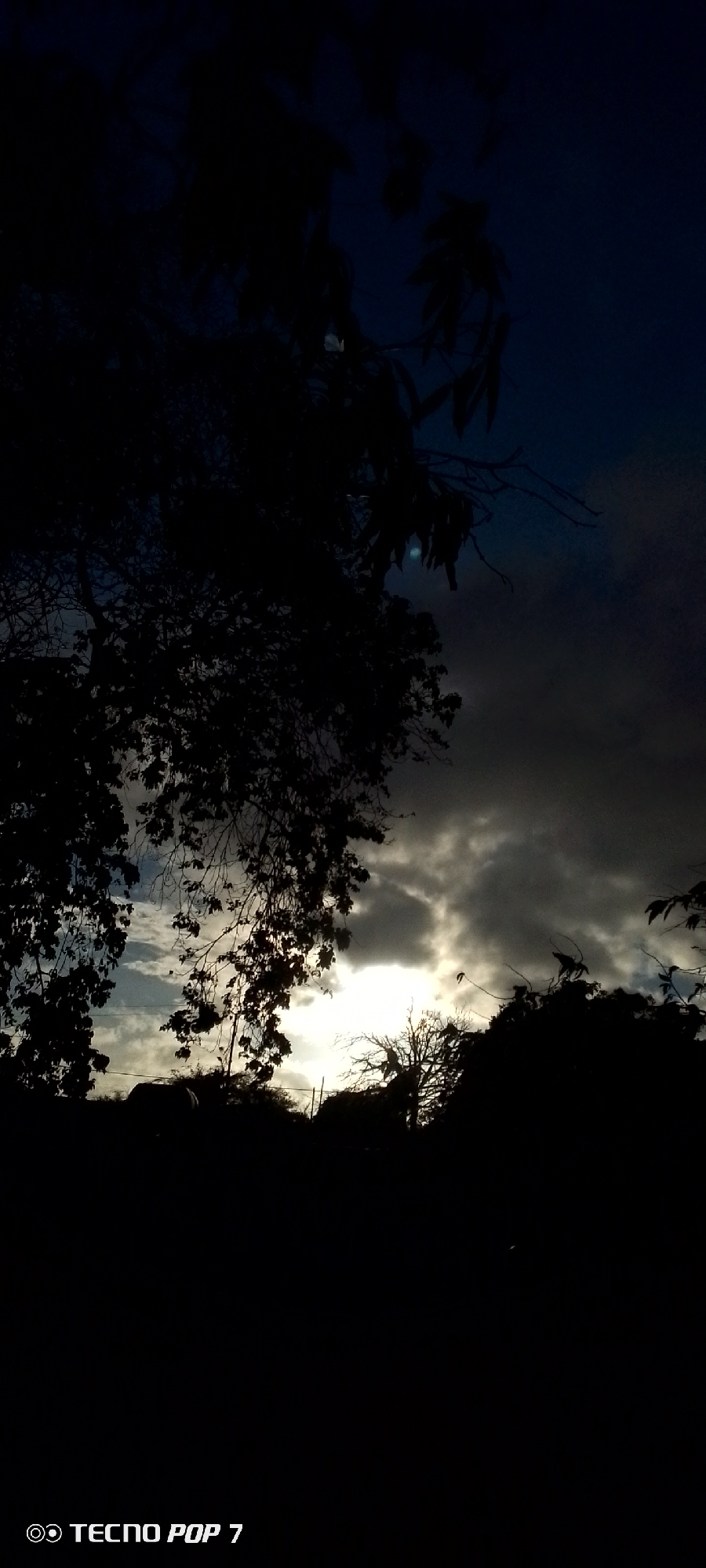 The image shows a dark scene with silhouettes of trees and foliage against a cloudy sky with a patch of bright light, possibly the sun, visible on the horizon. The sky transitions from dark blue at the top to lighter shades of gray and white near the horizon. The foreground is dark, obscuring details of the ground. The photograph was taken with a Tecno Pop 7 phone. The location is specified as Altagracia, Venezuela, but no location data or subjects in the image provide specific indicators.