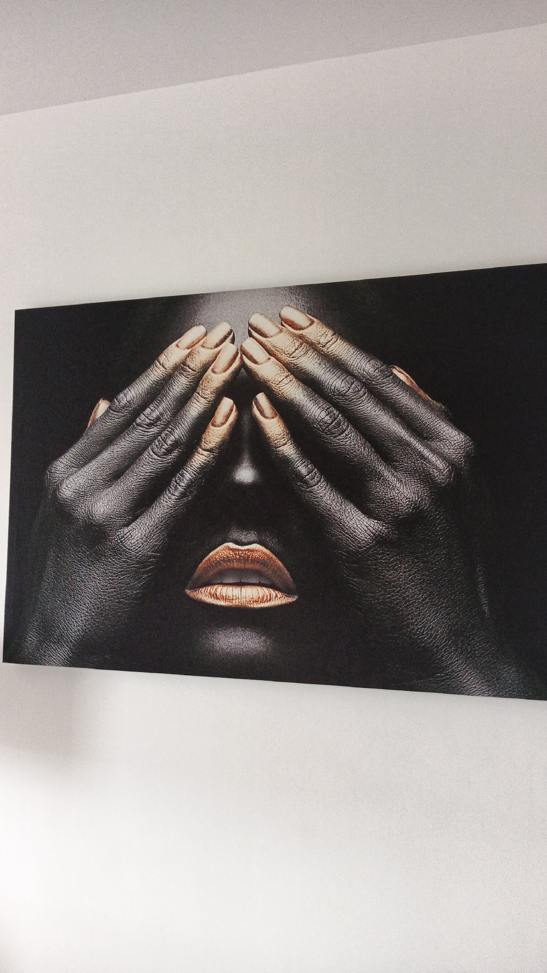 A close-up photograph of a painting features dark, textured skin with gold-tipped fingernails covering the eyes of a person. The lips, painted in a metallic gold hue, are visible below the hands. The background of the painting is black. The painting is mounted on a white wall, with a sliver of a white ceiling visible at the top of the frame. This artwork is located in Zielona Góra, Poland.