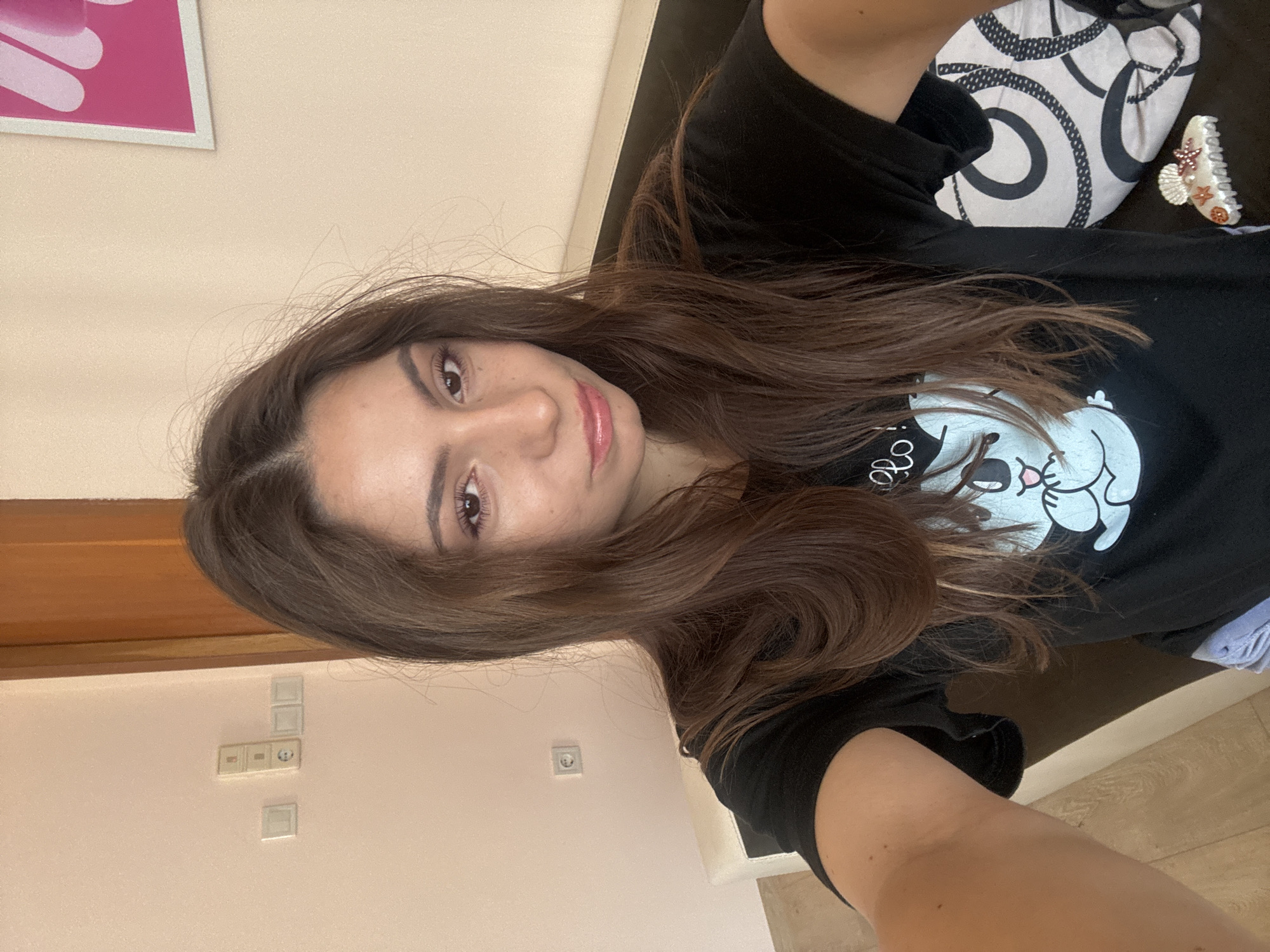 A young woman with long brown hair is taking a selfie indoors. She is wearing a black t-shirt with a cartoon character graphic on it. She is sitting on a couch with a pillow behind her. The background shows a light-colored wall with electrical outlets and a door. This appears to be a residential interior.