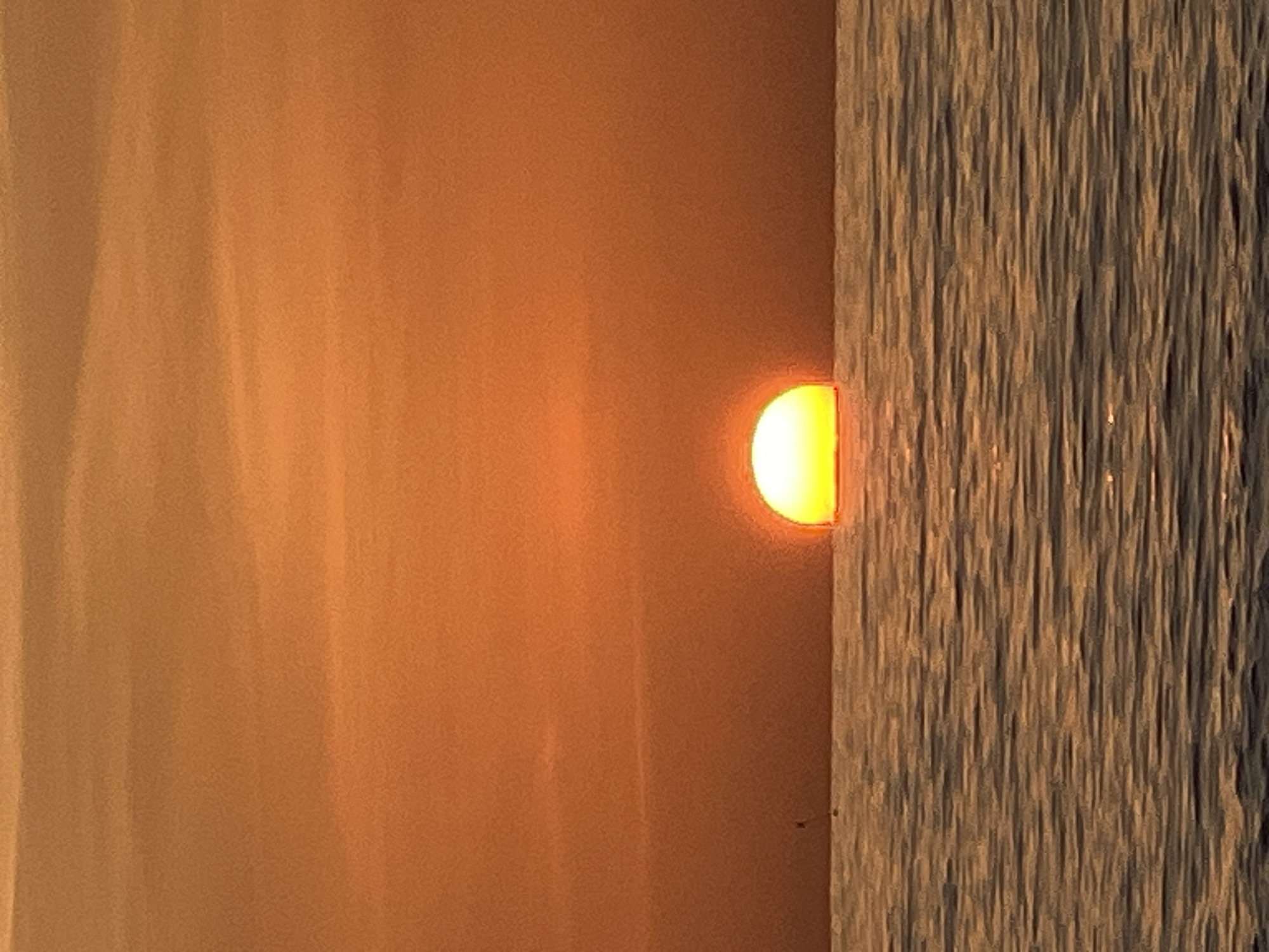 A half-sun is setting over a calm ocean, casting a warm orange glow on the sky. The sun's lower half is submerged below the horizon, while its upper half shines brightly. The water's surface reflects the sunset's colors in rippling patterns. In the distance, a faint silhouette of a small boat or person is visible on the water. The scene suggests a peaceful evening at a coastal location.