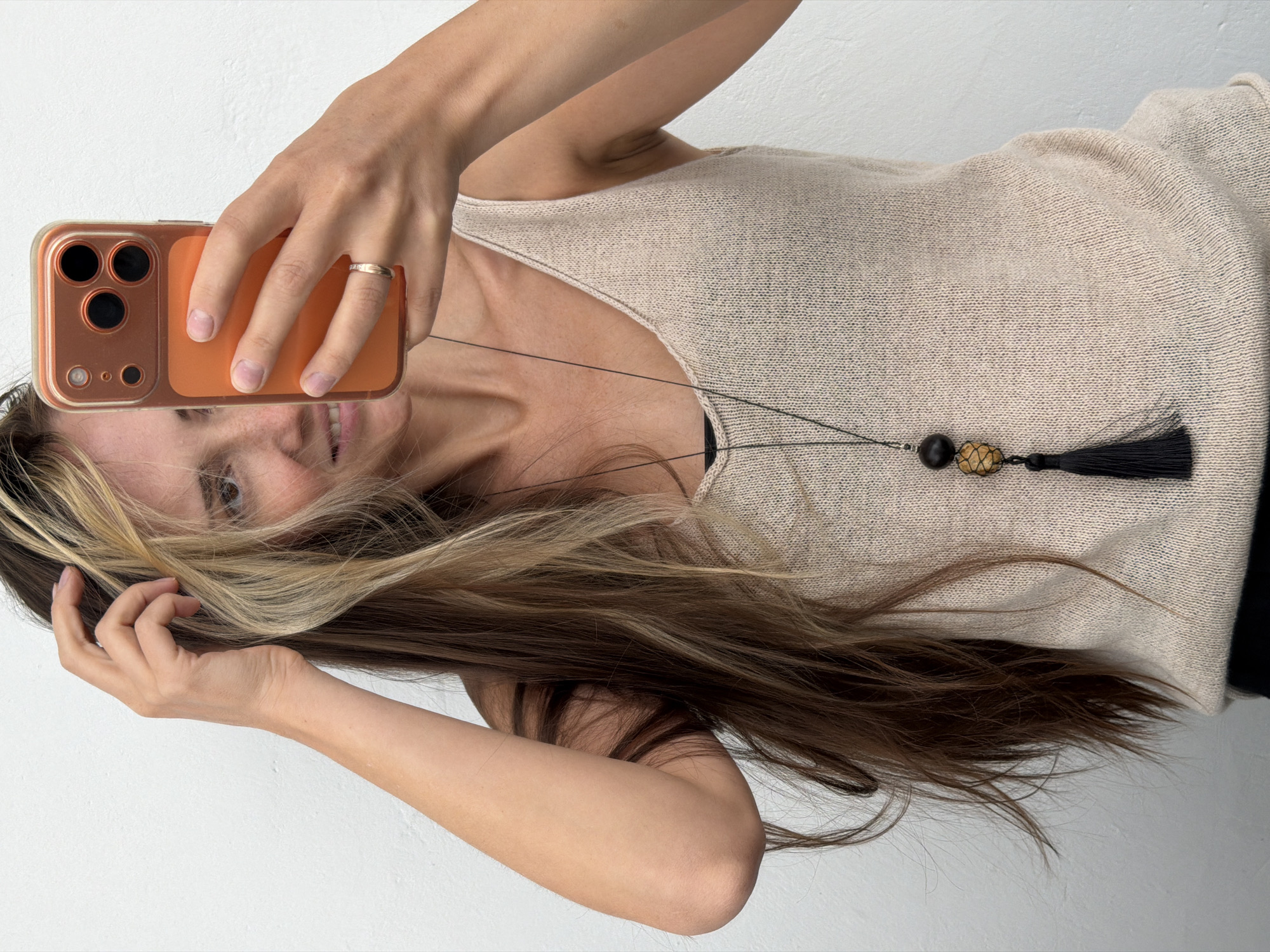 A woman with long, multi-toned brown hair is taking a selfie in front of a plain white wall. She is wearing a light beige, knitted tank top and a long necklace with dark beads and a black tassel. Her right hand is raised, touching her hair, while her left hand holds an orange smartphone. A ring is visible on her left ring finger. The background is a simple, unadorned white wall, typical of indoor settings in urban environments, such as apartment complexes or studios. There are no indications of outdoor elements or specific landmarks that would place this scene in Khimki, Russia. The lighting is even, suggesting artificial light or diffused natural light.