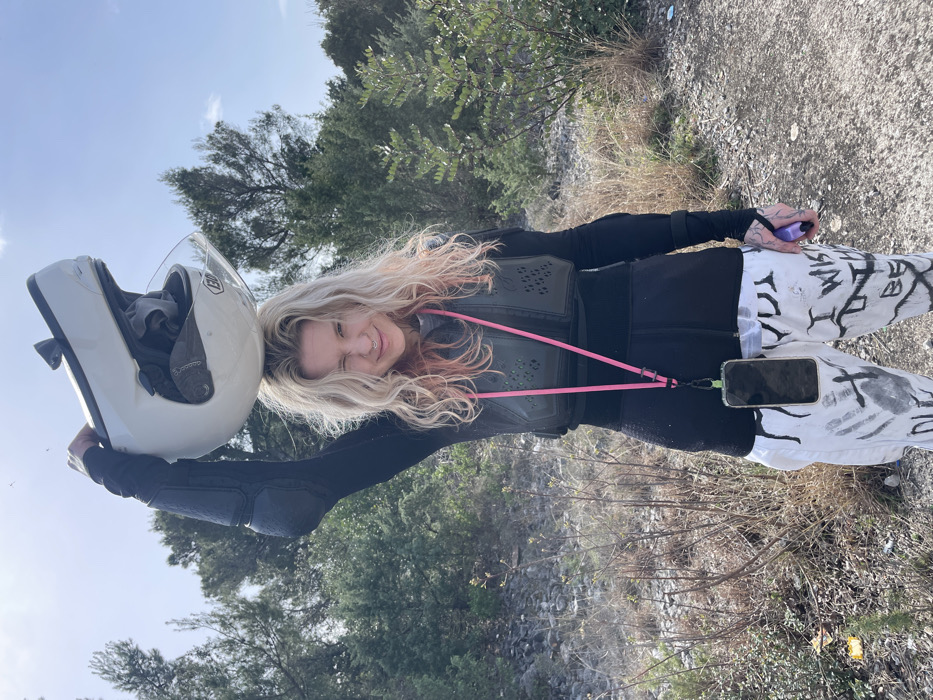 An individual with wavy blonde hair, light pink streaks, and a nose piercing stands outdoors in Bigova, Montenegro, under a clear blue sky. They face the camera, winking their right eye and smiling slightly. Their left arm is raised, holding a white Shoei full-face motorcycle helmet above their head. They are dressed in a black long-sleeved top with visible elbow pads, overlaid by a black, perforated chest protector. A pink lanyard hangs around their neck, suspending a black smartphone in a clear case at waist level. They wear white pants adorned with black text and handprint-like designs. A purple object is partially visible in their right hand. Tattoos are present on their left hand and wrist. The background consists of sparse vegetation including green bushes and small trees, with a ground composed of dirt and gravel.
