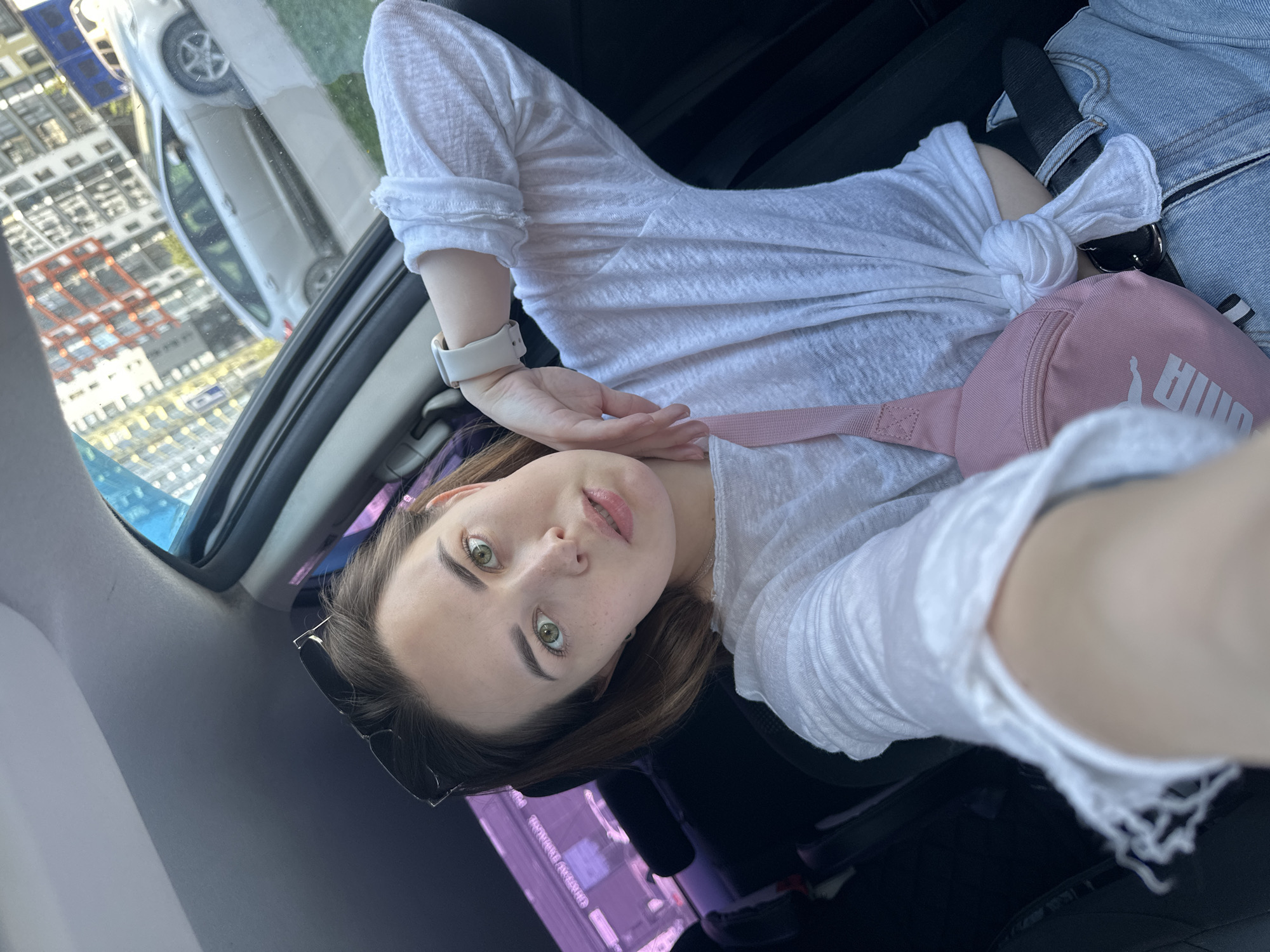 A young woman with green eyes is lying down in a car, taking a selfie. She is wearing a white long-sleeve shirt tied at the waist, a white smartwatch, and a light pink Puma fanny pack. Sunglasses are tucked into her hair. The car appears to be parked on a street in Perm, Russia, as seen through the window. The buildings reflect in the car window.