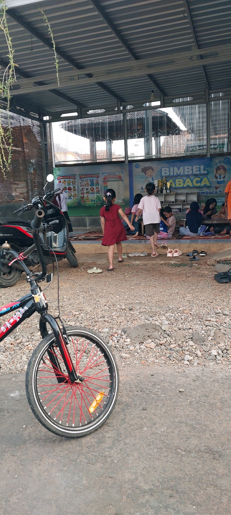 The image depicts an outdoor scene in Keling, Indonesia, likely a community learning center or outdoor classroom. In the foreground, a bicycle with a black frame and bright red spokes is positioned diagonally, its front wheel prominently featured. Behind the bicycle, a black motorcycle is parked. The ground is covered with gravel and some asphalt.
In the mid-ground, several children and adults are gathered. A young girl in a red dress is walking away from the camera. Another child, wearing a pink shirt and brown shorts, is walking alongside her. Several other children are seated on mats on the ground, some of them engaged in conversation. Behind the children, a mural-style poster with Indonesian text and cartoon characters is displayed on a wall, likely advertising educational programs or events. A shelf unit with various items, including trophies, is visible against the wall.
The background shows the exterior of a building with a corrugated metal roof supported by a metal frame. A woven bamboo screen is visible to the left. The overall environment suggests a somewhat informal, community-oriented setting.