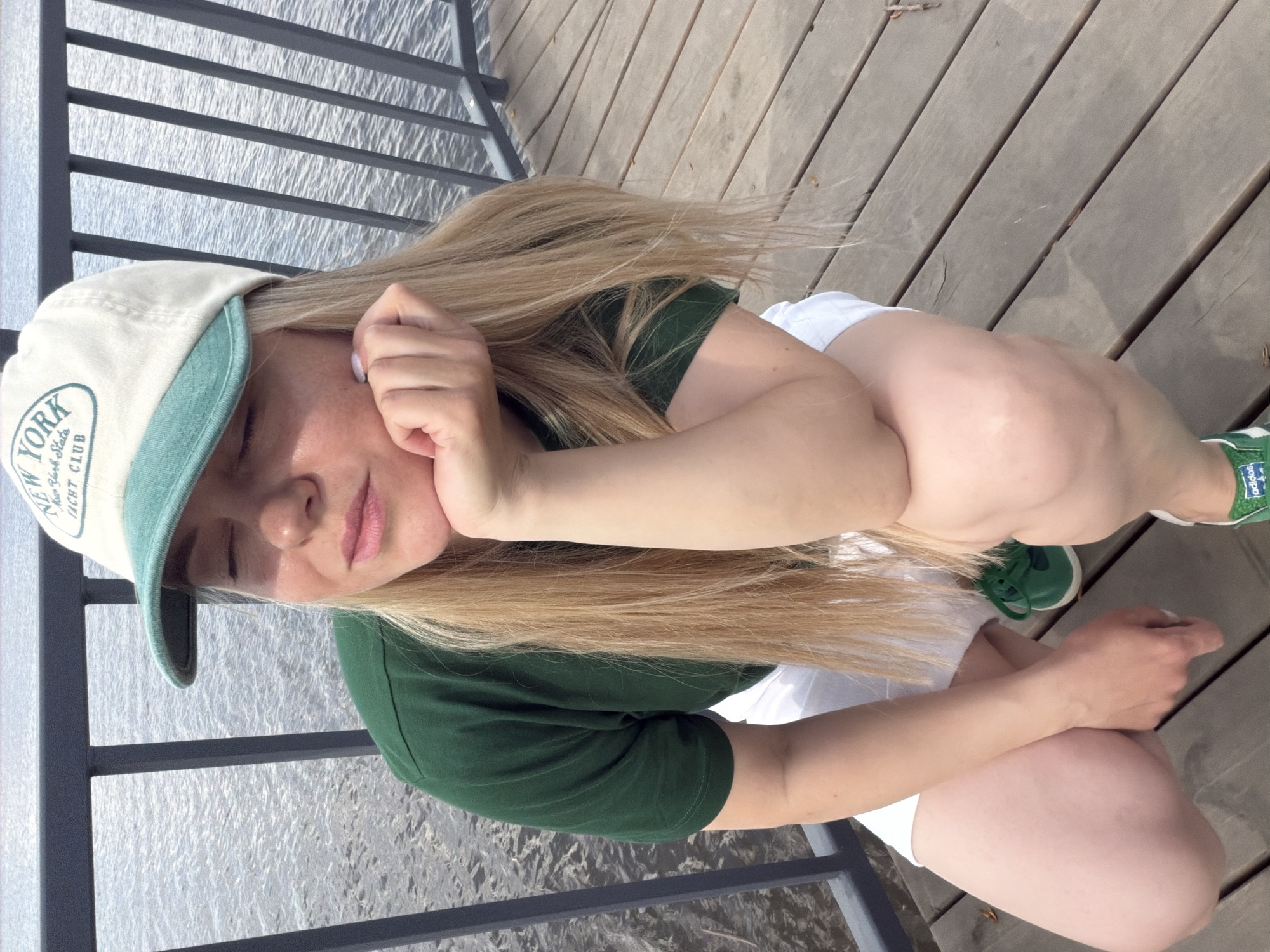A young woman is crouching on a wooden deck near a body of water in Votkinsk, Russia. She is wearing a green t-shirt, white shorts, and green Adidas sneakers. She has a light-colored baseball cap with "NEW YORK YACHT CLUB" printed on it. She is resting her face in her hand, with her eyes closed. A metal railing separates the deck from the water.